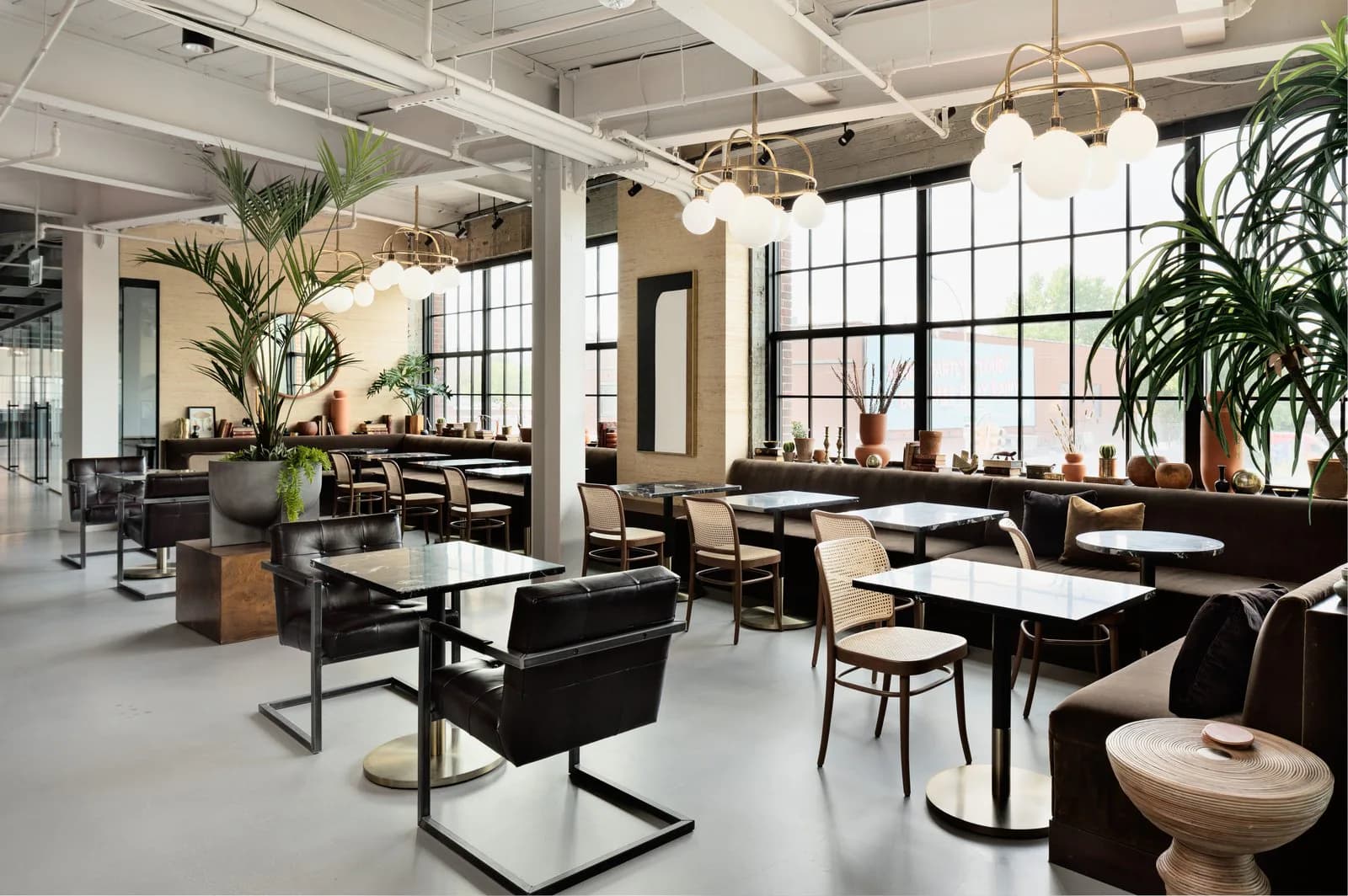 Modern office lounge with black leather seating, natural wood tables, abundant greenery, industrial ceiling, and large windows overlooking city views by Elide Rathborne.