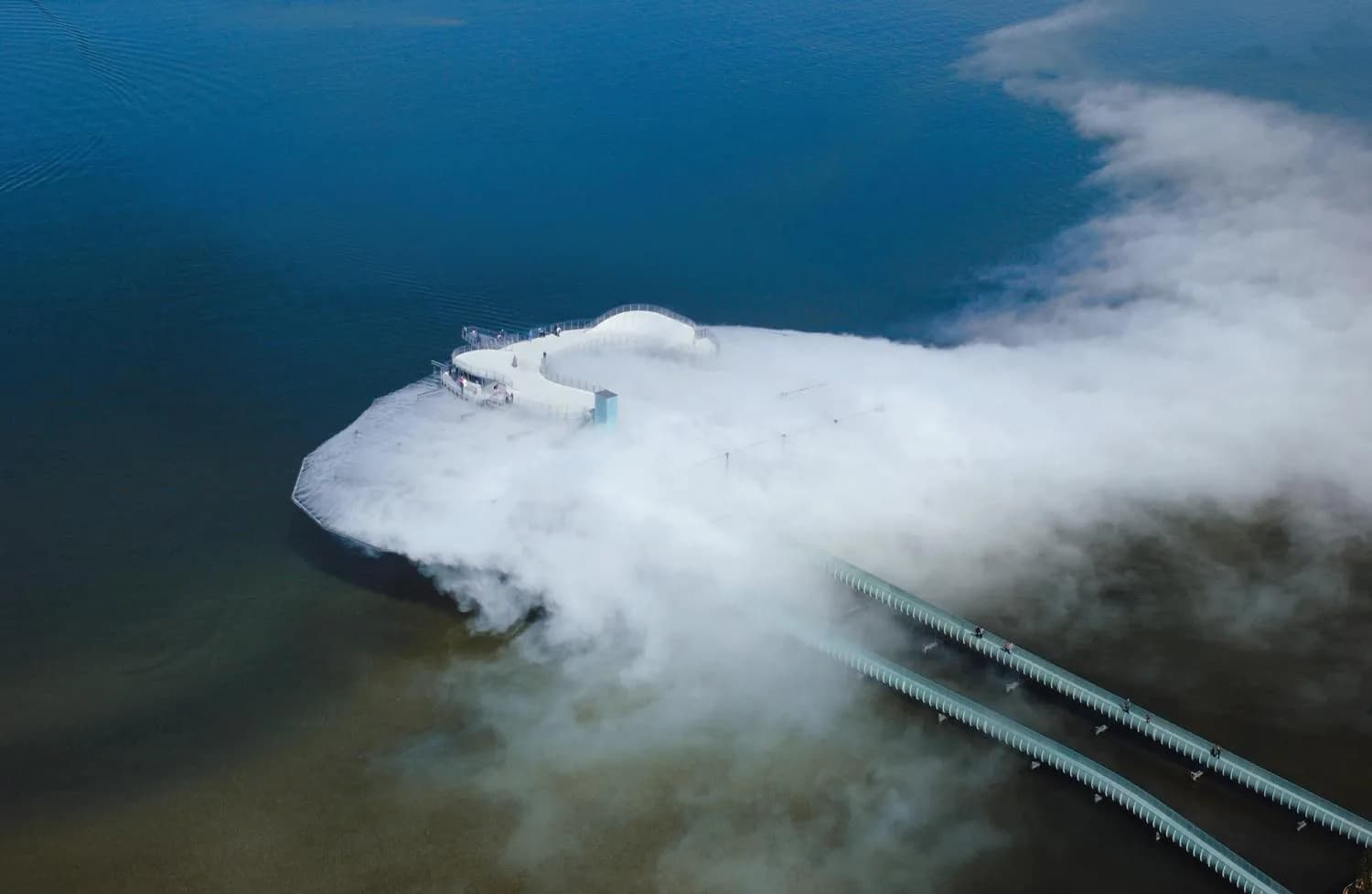 Aerial view of a white architectural structure emerging from mist over water with green piping visible, designed by Elizabeth Diller.