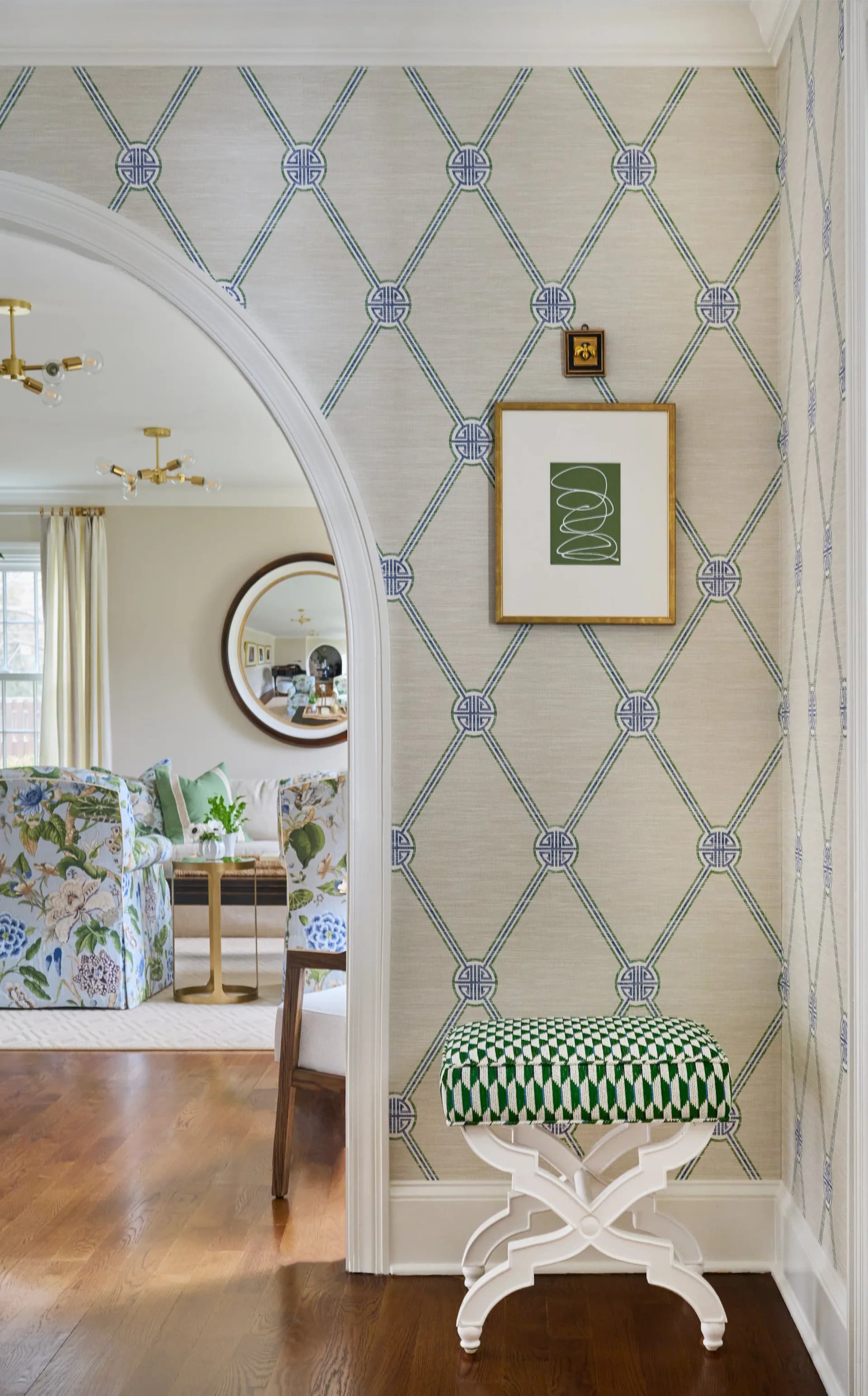 Elegant hallway with geometric patterned wallpaper, white curved archway, round mirror, framed botanical print, and green patterned bench by Ellen Marment.
