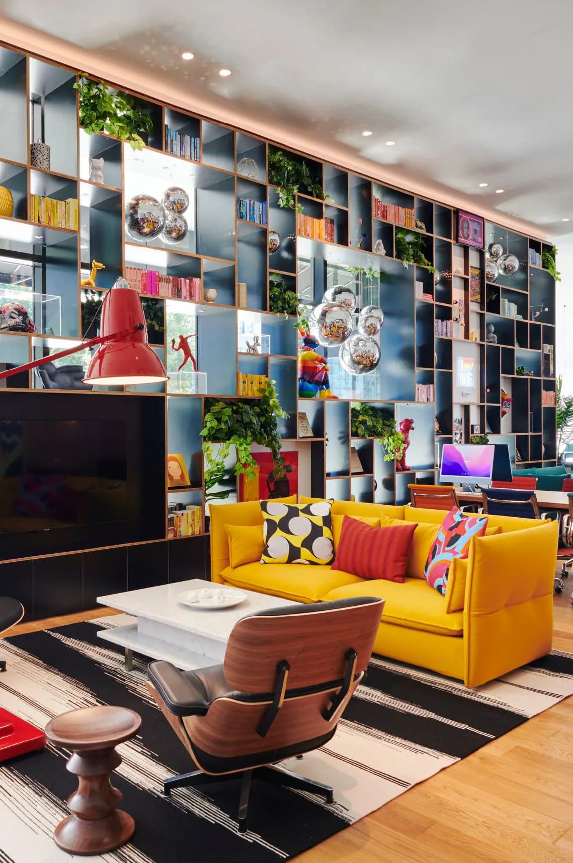 Colorful living room interior with yellow sofa, geometric patterned rug, red pendant lamp, and floor-to-ceiling modular shelving with plants and decorative objects by Ellen Vega.