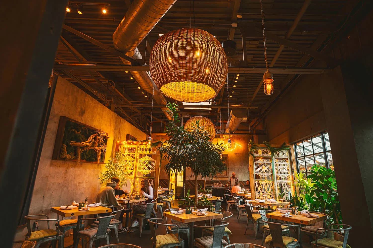 Warm-lit restaurant interior with woven pendant light, exposed ceiling, lush greenery, and wooden tables arranged throughout by Emad Hasan.
