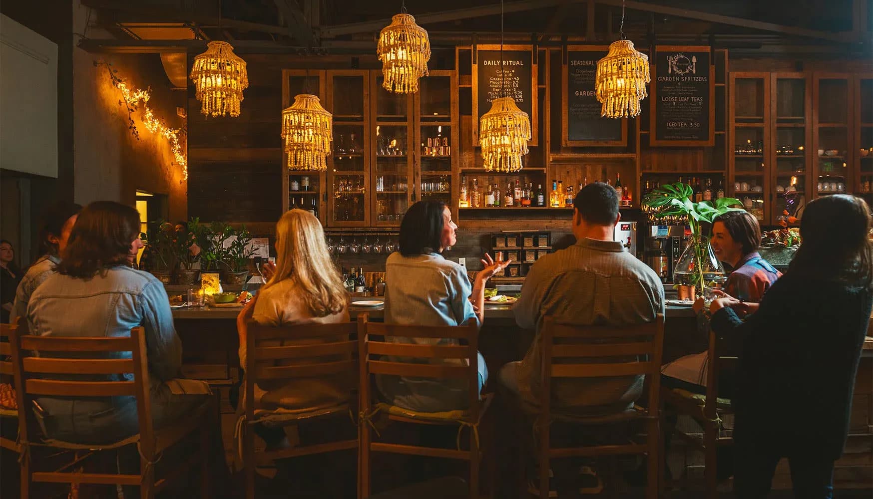 Warm-toned bar interior with wooden cabinetry, golden pendant lights, and customers seated at the counter, designed by Emad Hasan.