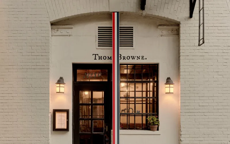 Storefront entrance for Thom Browne featuring dark wood doors with gridded windows, cream curved archway, flanking sconces, and red vertical striping accent by Emily Paulson.