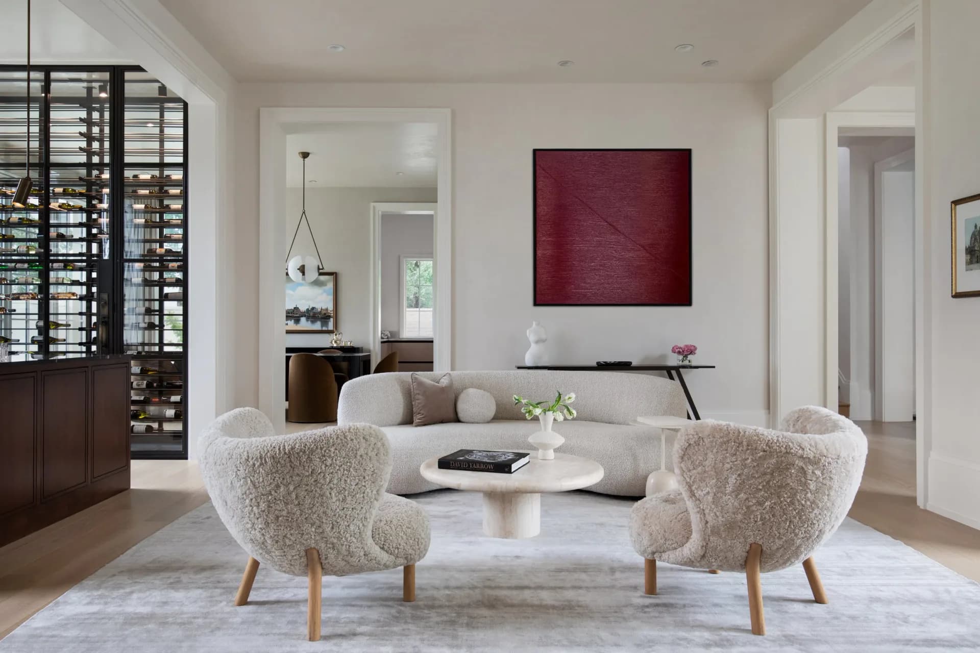 Modern living room with cream upholstered chairs, white marble table, burgundy artwork, and dark wood shutters, designed by Emma Corner.