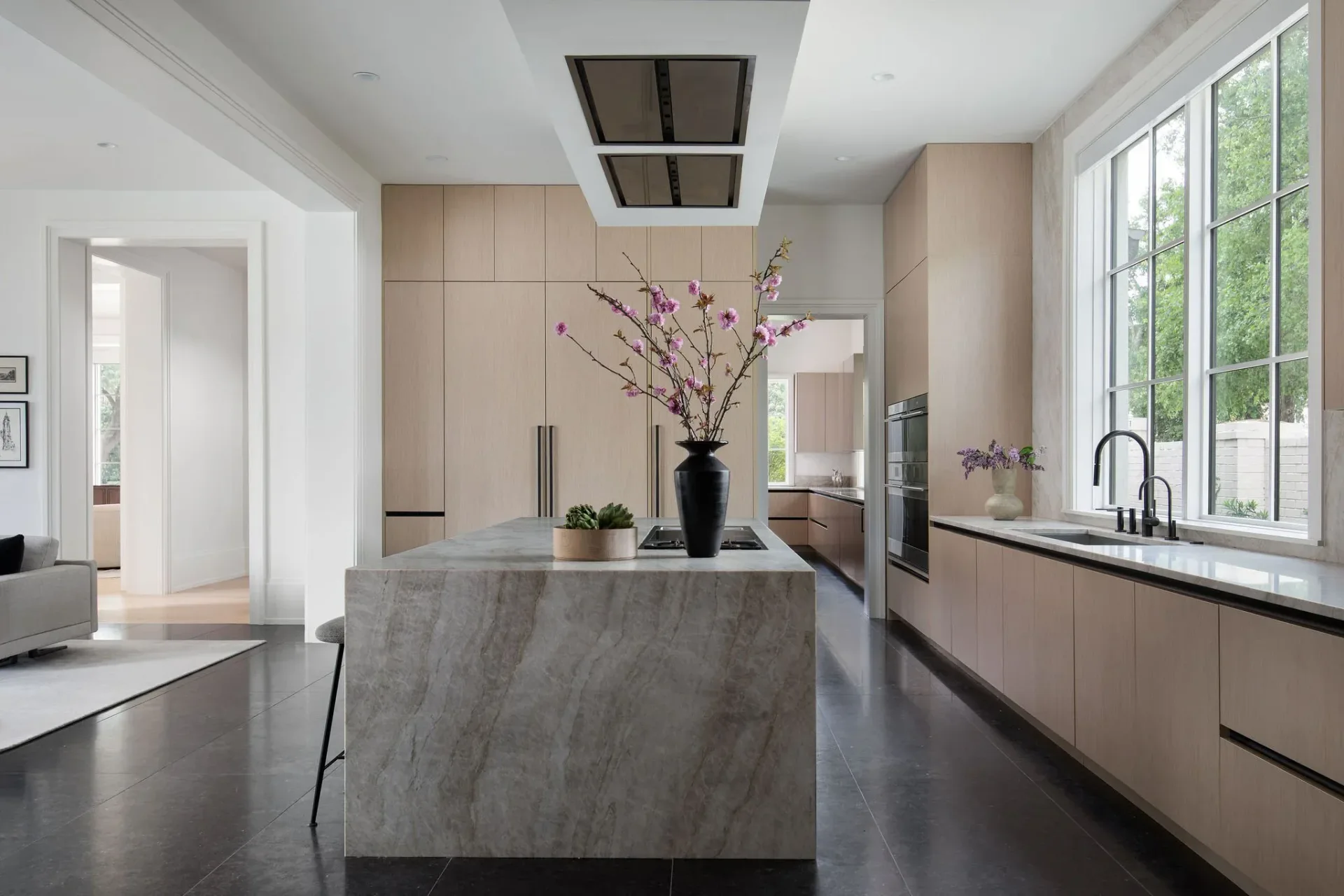 Modern kitchen with light wood cabinetry, gray marble island, black countertops, and large windows, designed by Emma Corner.