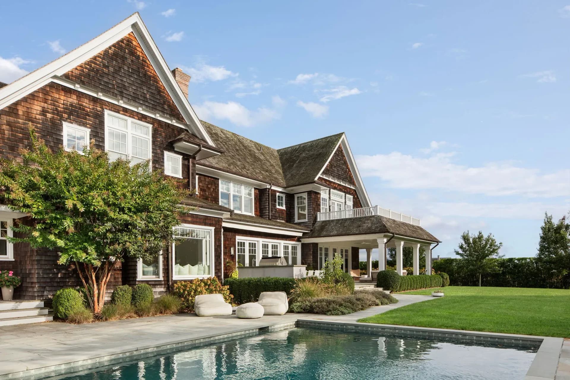 Modern residential architecture featuring a dark shingle-clad farmhouse with stone accents, manicured landscaping, and an elegant poolside design by Emma Corner.
