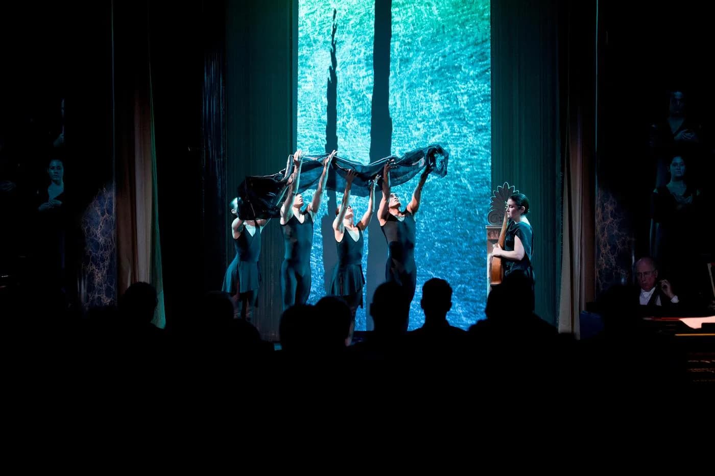 Theater stage with dramatic turquoise backdrop and performers suspended mid-air in dynamic poses, audience silhouetted in foreground, theatrical lighting design by Enrique Pincay.