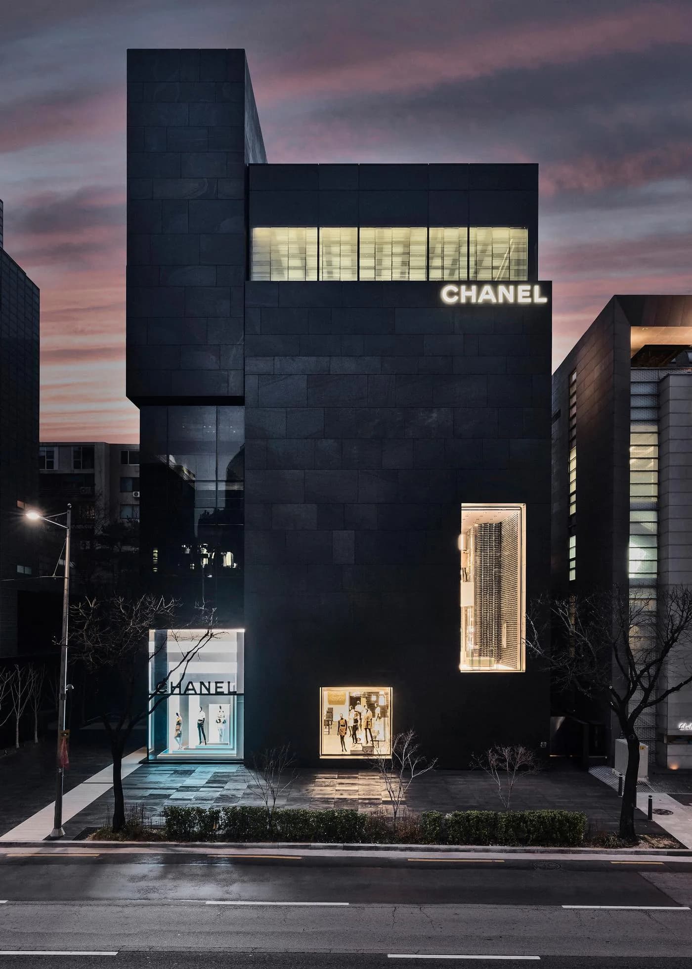 A modern black architectural facade for a Chanel boutique with illuminated storefront windows and golden interior lighting at dusk, designed by Enrique Pincay.
