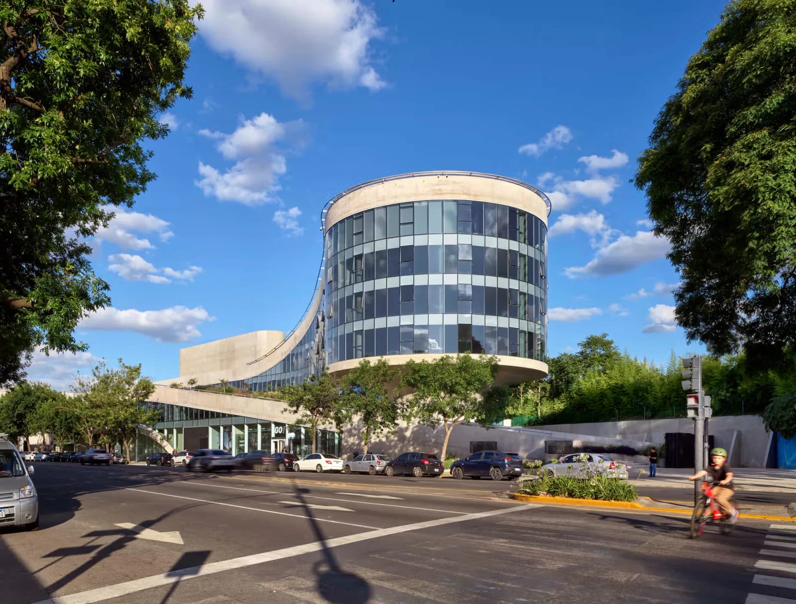 Curved modernist cylindrical building with horizontal banding, glass windows, and curved base structure set in urban plaza with trees by Eran Chen.