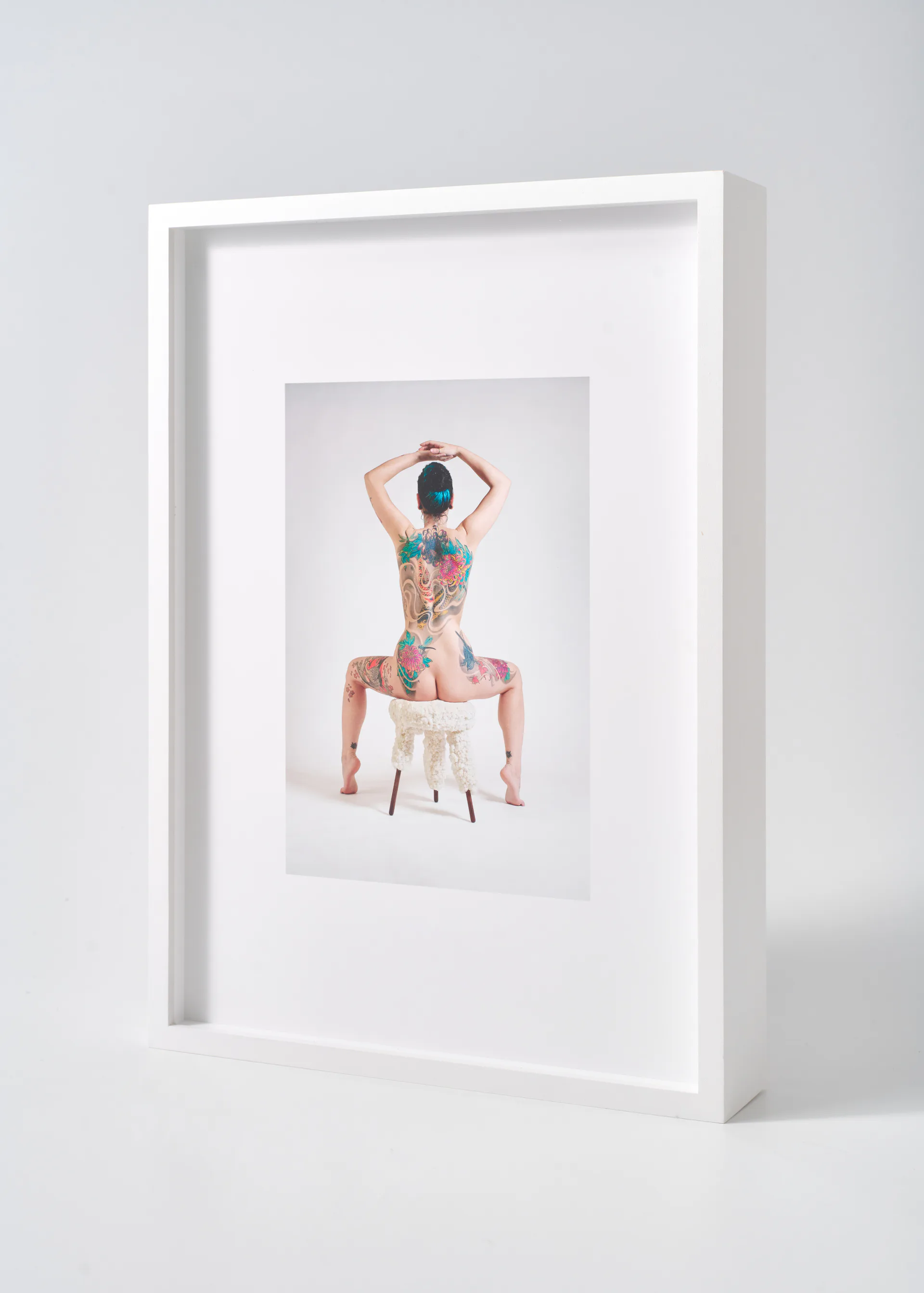 White framed art installation displaying a surreal portrait of a figure with colorful patterned body art sitting on a white chair, by Erez Nevi Pana.
