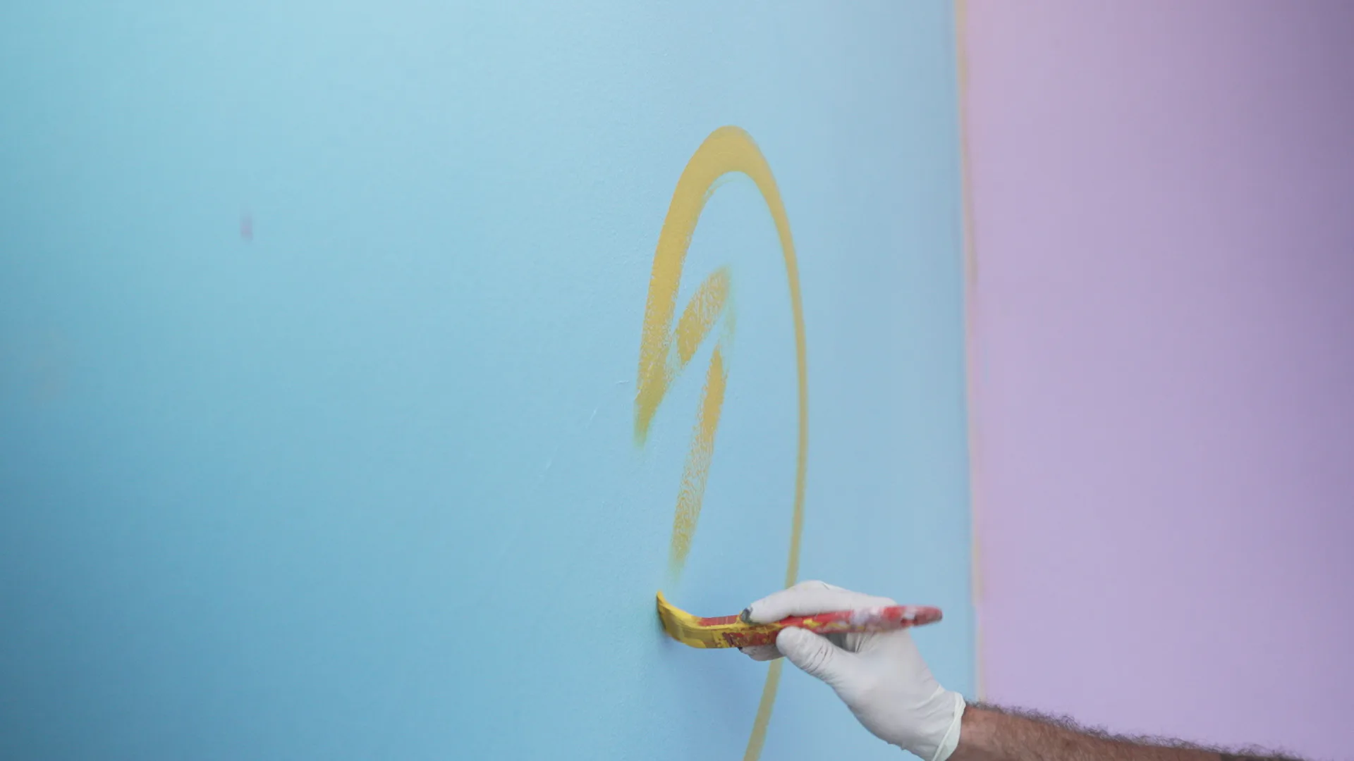 Hand applying yellow paint with brush to blue and pink gradient wall, creating curved brushstroke design by Erez Nevi Pana.