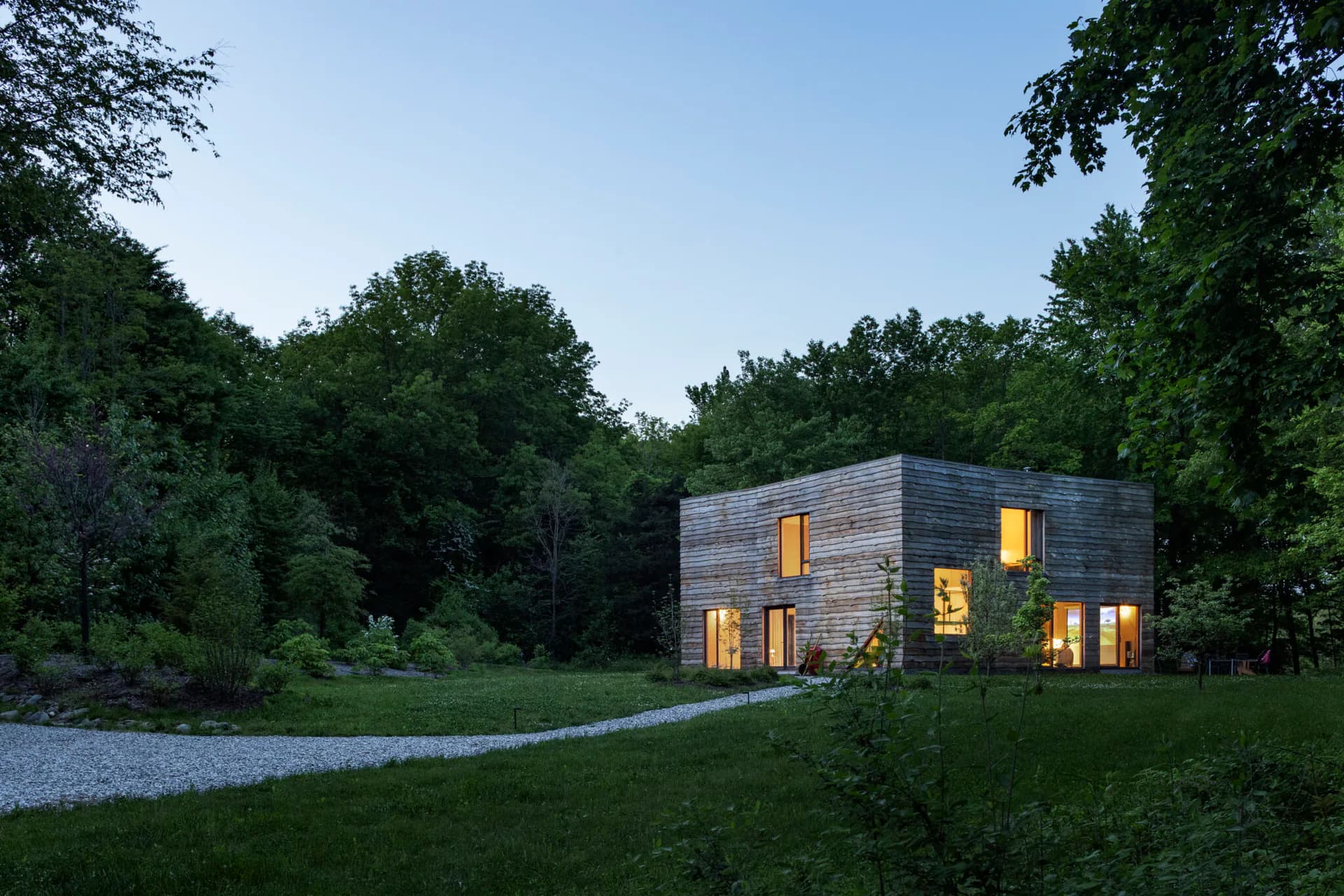 Modern cubic residence with charred wood exterior and warm-lit windows nestled in a verdant forest landscape by Eric Bunge.