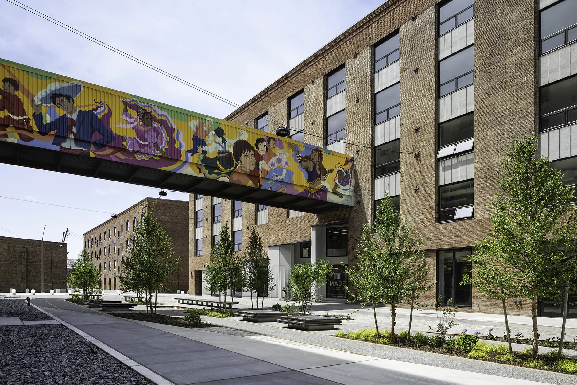Contemporary urban plaza with brick buildings, colorful mural bridge, young trees, and paved courtyard by Eric Bunge.