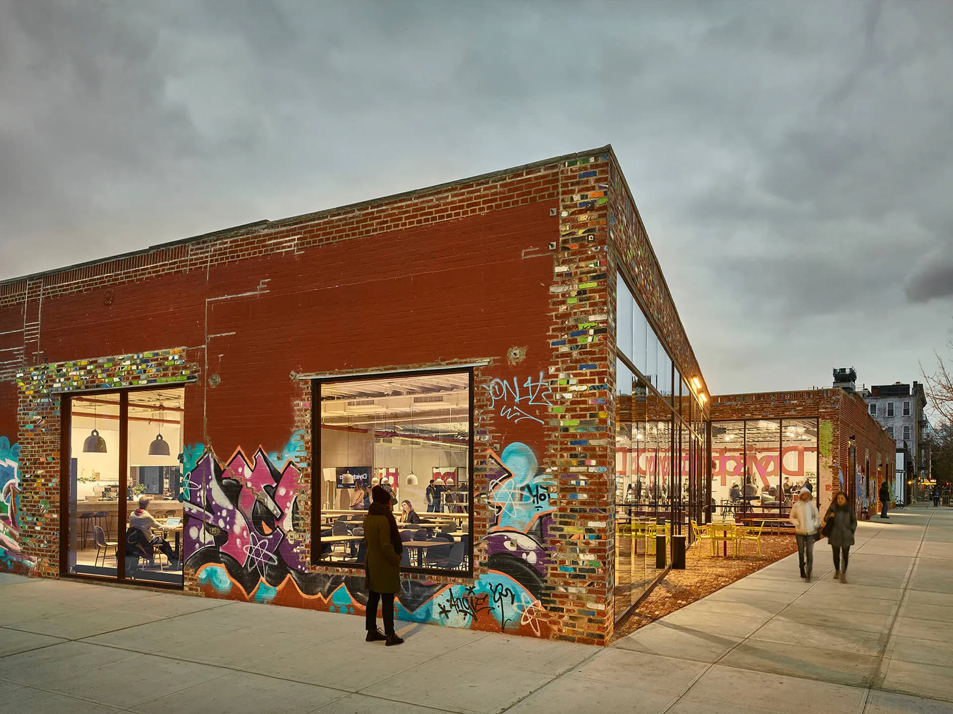 Brick corner building with red facade and colorful street art murals, large storefront windows, and pedestrians on plaza by Eric Bunge.