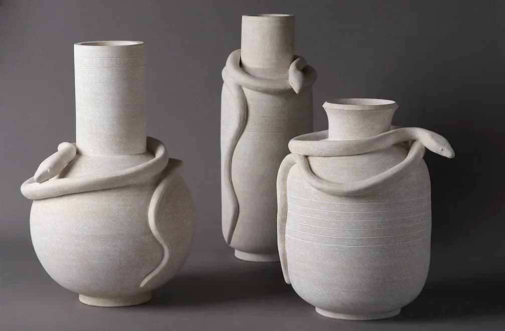 Three white ceramic vessels with organic sculptural forms and curved handles, displayed against a gray background by Eric Roinestad.