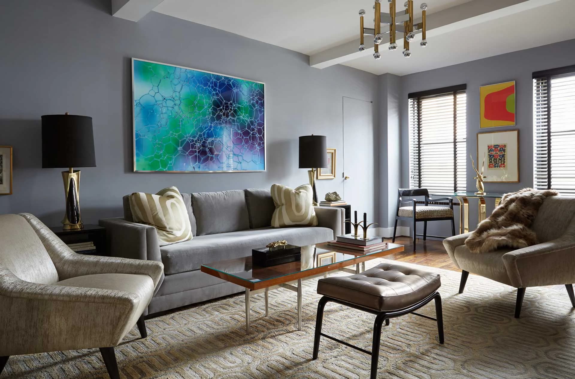 Contemporary living room with gray sofa, brass accents, and blue abstract artwork, featuring geometric coffee tables and brass pendant lighting by Eric Winnick.