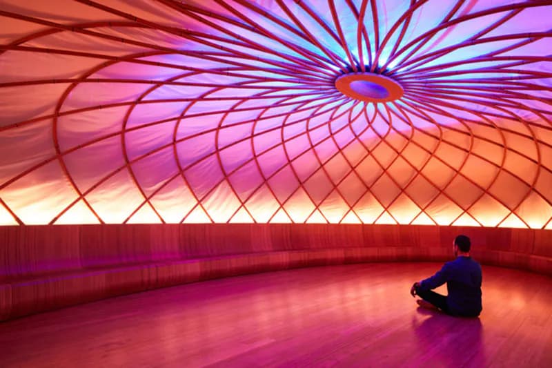 A person sits in a luminous pink and magenta interior installation with radiating geometric ceiling patterns and glowing fabric walls by Erik Tietz.