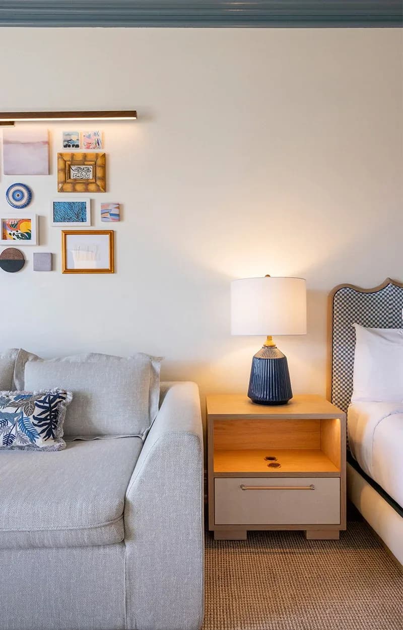 Modern bedroom corner with gray upholstered sofa, wooden nightstand with white lamp, checkered headboard, and gallery wall of framed artwork and decorative plates by Erika Dray.