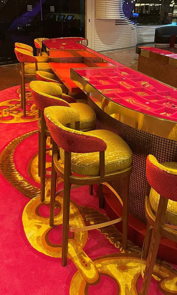 Modern bar interior with vibrant red and gold color palette, featuring curved yellow upholstered bar stools and matching red leather seating, geometric patterned flooring, and sleek wood-framed furniture by Erika Dray.