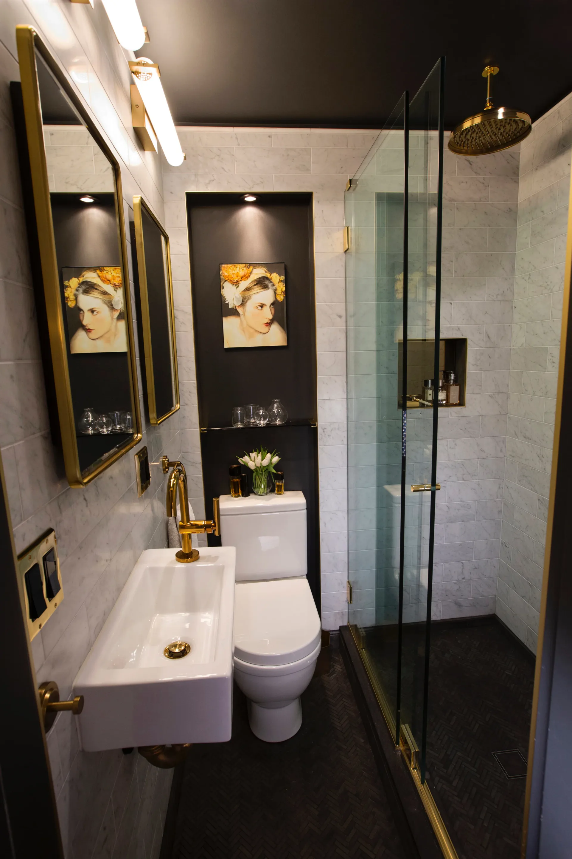 Luxe bathroom with black accent wall, gold-framed mirrors, white fixtures, marble tile, and glass shower enclosure by Erika Schmitt.