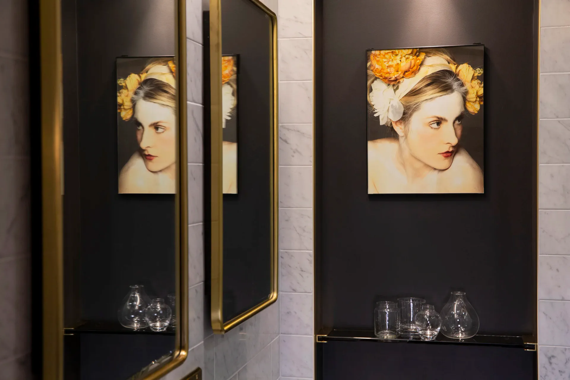 Dark gallery wall with gold-framed mirror and two portrait artworks featuring golden floral elements, styled display shelves below by Erika Schmitt.