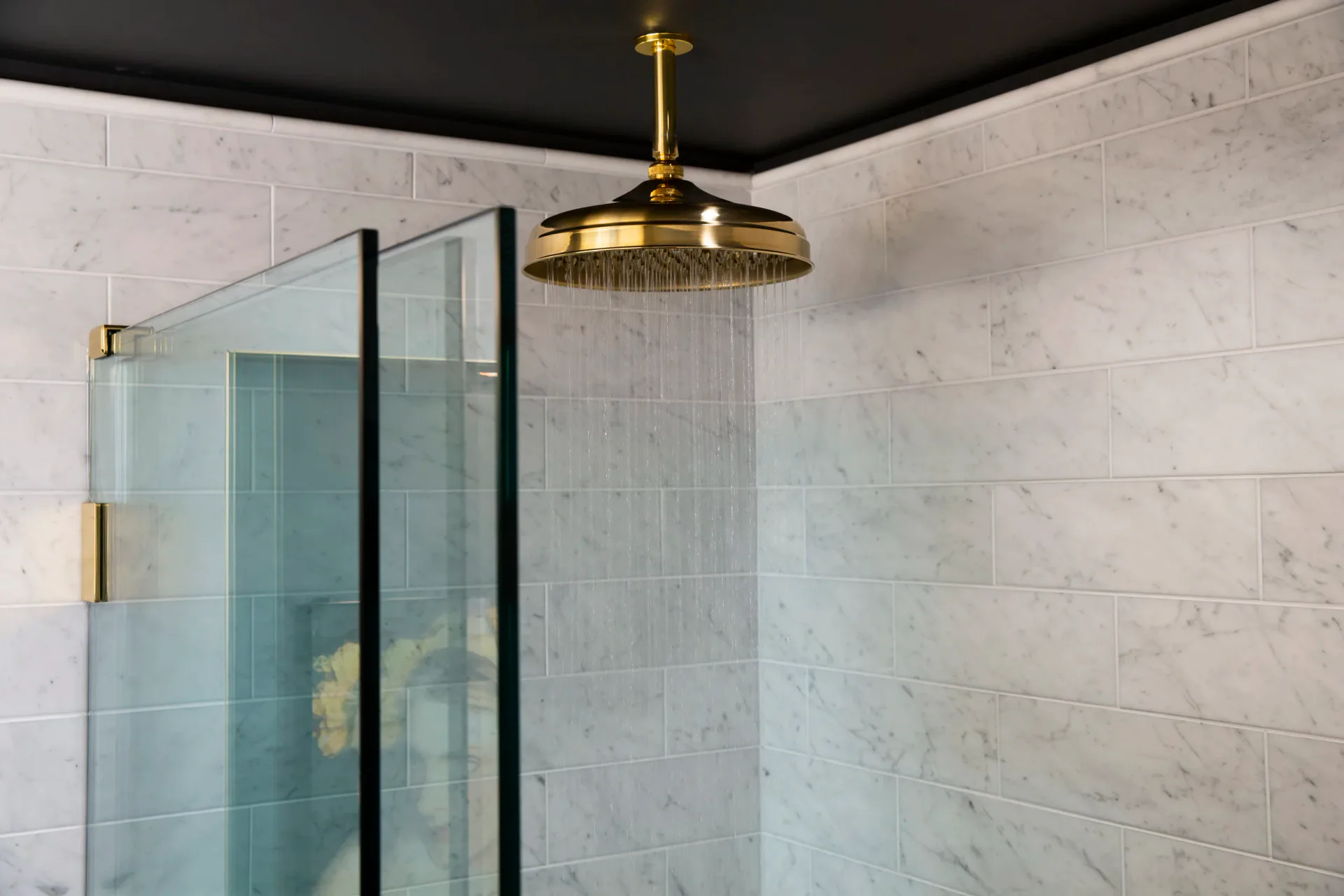 Modern bathroom with marble tile walls, black-framed glass shower enclosure, and brass rainfall showerhead by Erika Schmitt.