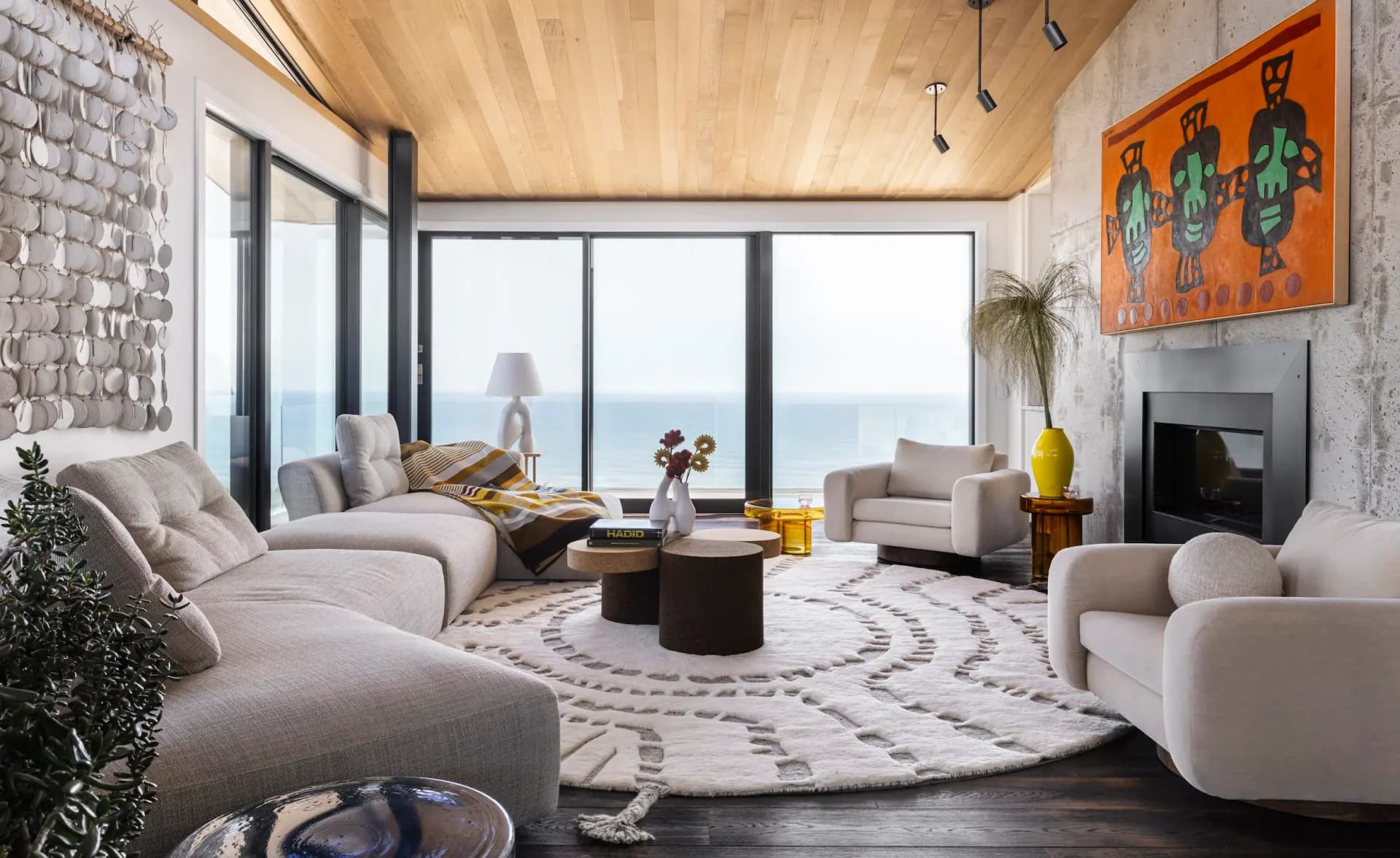 Modern luxury living room with curved cream furniture, wooden ceiling, ocean views, patterned area rug, and fireplace by Erin Fearins.