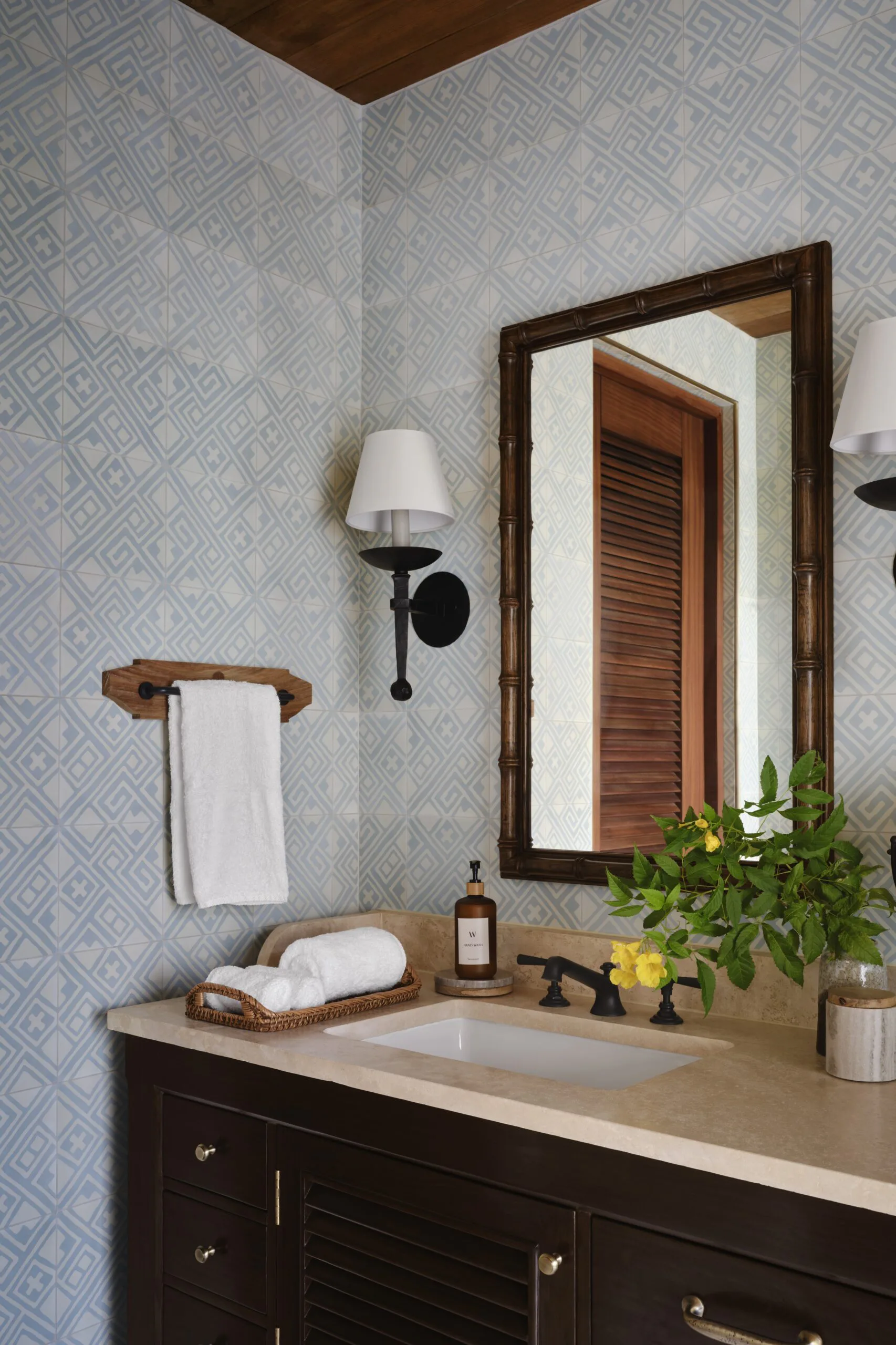 Bathroom vanity with dark wood cabinetry, light countertop, framed mirror, geometric patterned wallpaper, and coordinating fixtures by Ernesto Gloria.