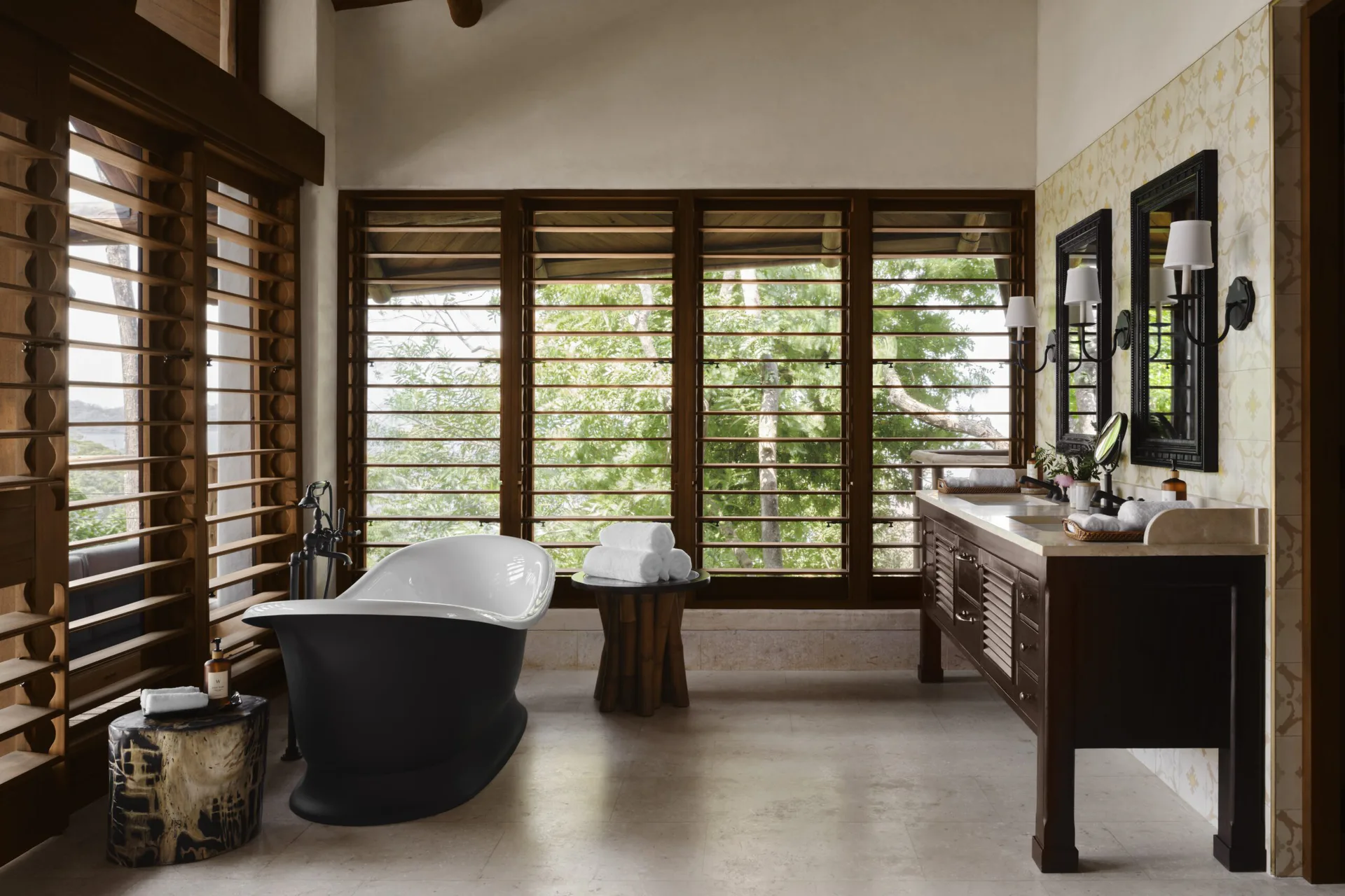 Luxurious spa bathroom with black freestanding bathtub, dark wood vanity, louvered windows, concrete flooring, and natural materials by Ernesto Gloria.