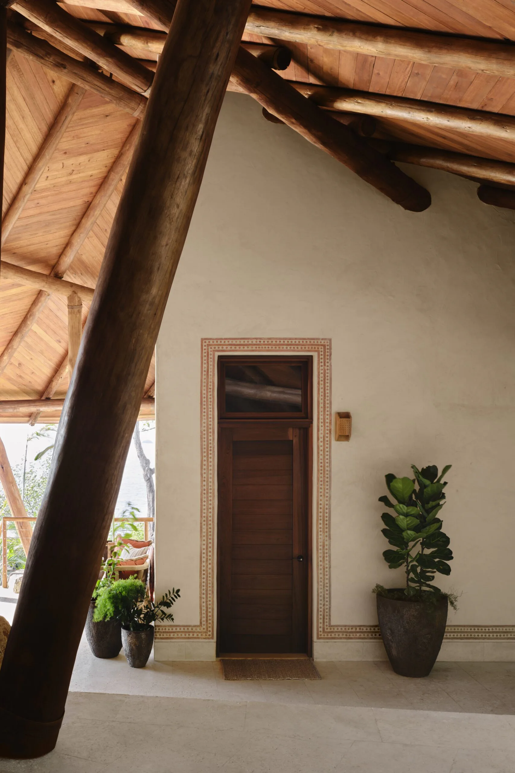 Minimalist interior with exposed wooden beam ceiling, cream walls, dark wooden door with decorative frame, and potted plants by Ernesto Gloria.