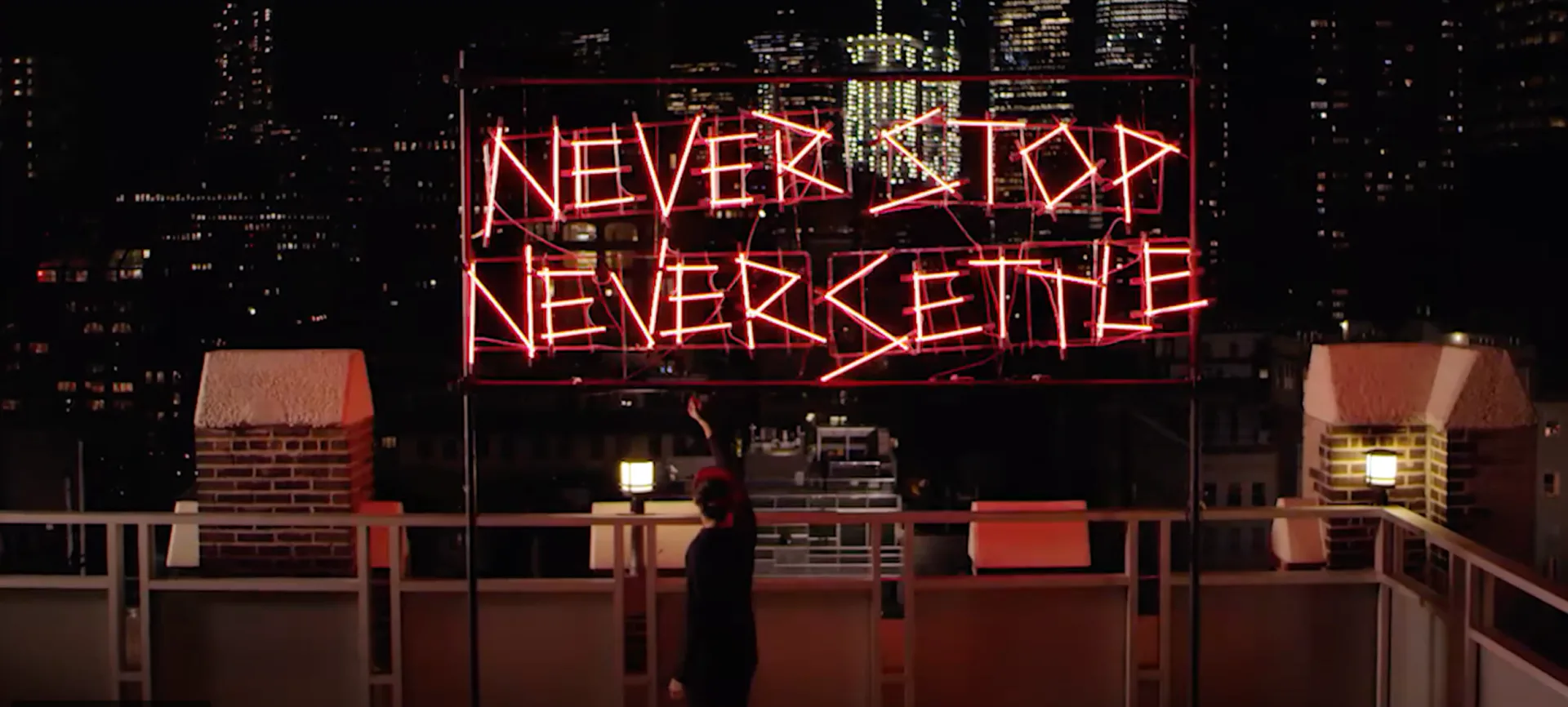 Rooftop installation with neon signage reading 'NEVER STOP NEVER SETTLE' against Manhattan skyline, featuring red architectural elements and seating by Esteban Salazar-Cucalon.