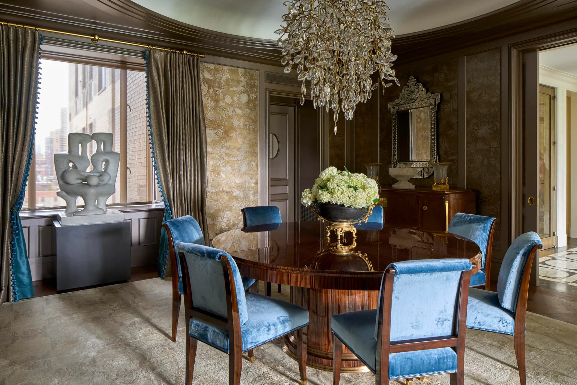 Elegant formal dining room with dark wood paneling, blue upholstered chairs, crystal chandelier, and white floral centerpiece by Evan Ainbinder.