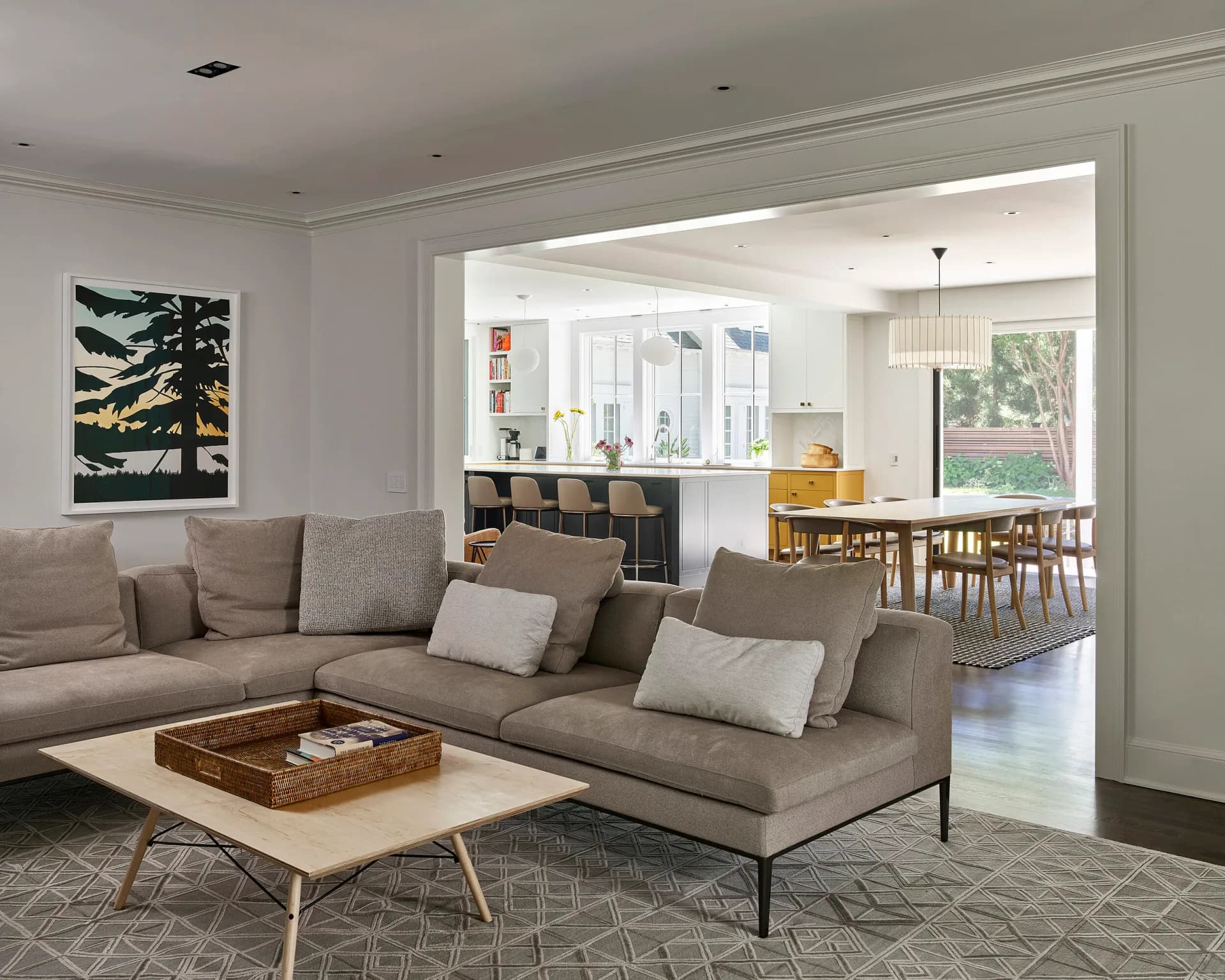 Modern living room with gray sectional sofa, light wood coffee table, and view into kitchen and dining area with natural wood furniture by Eve Wildrick.