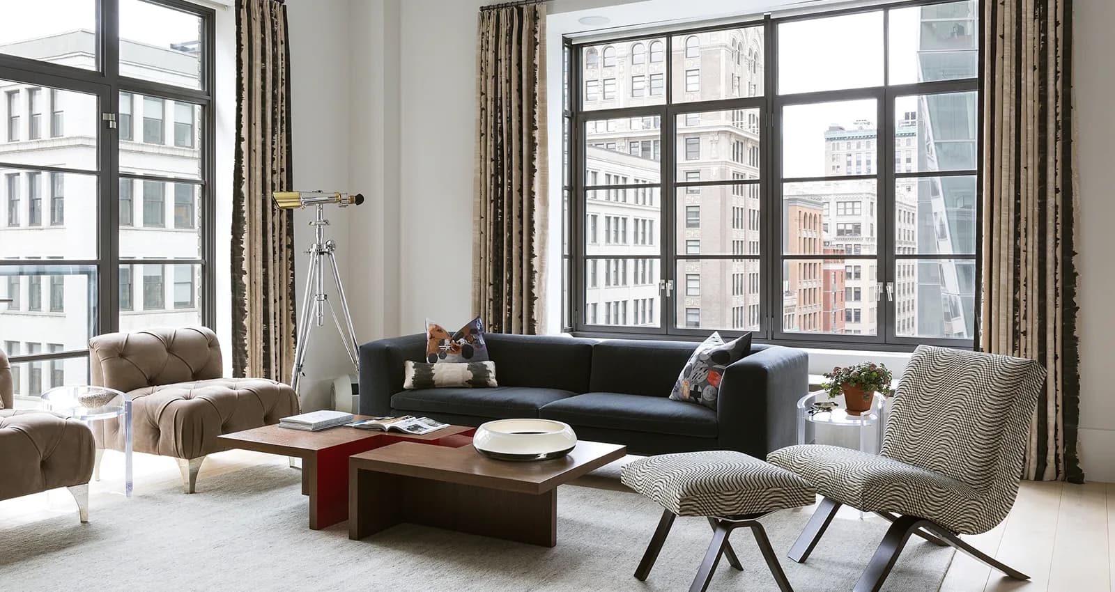 Modern living room with floor-to-ceiling windows overlooking city buildings, featuring a dark sofa, tan armchairs, wooden coffee table, and patterned accent chair by Eve Wildrick.