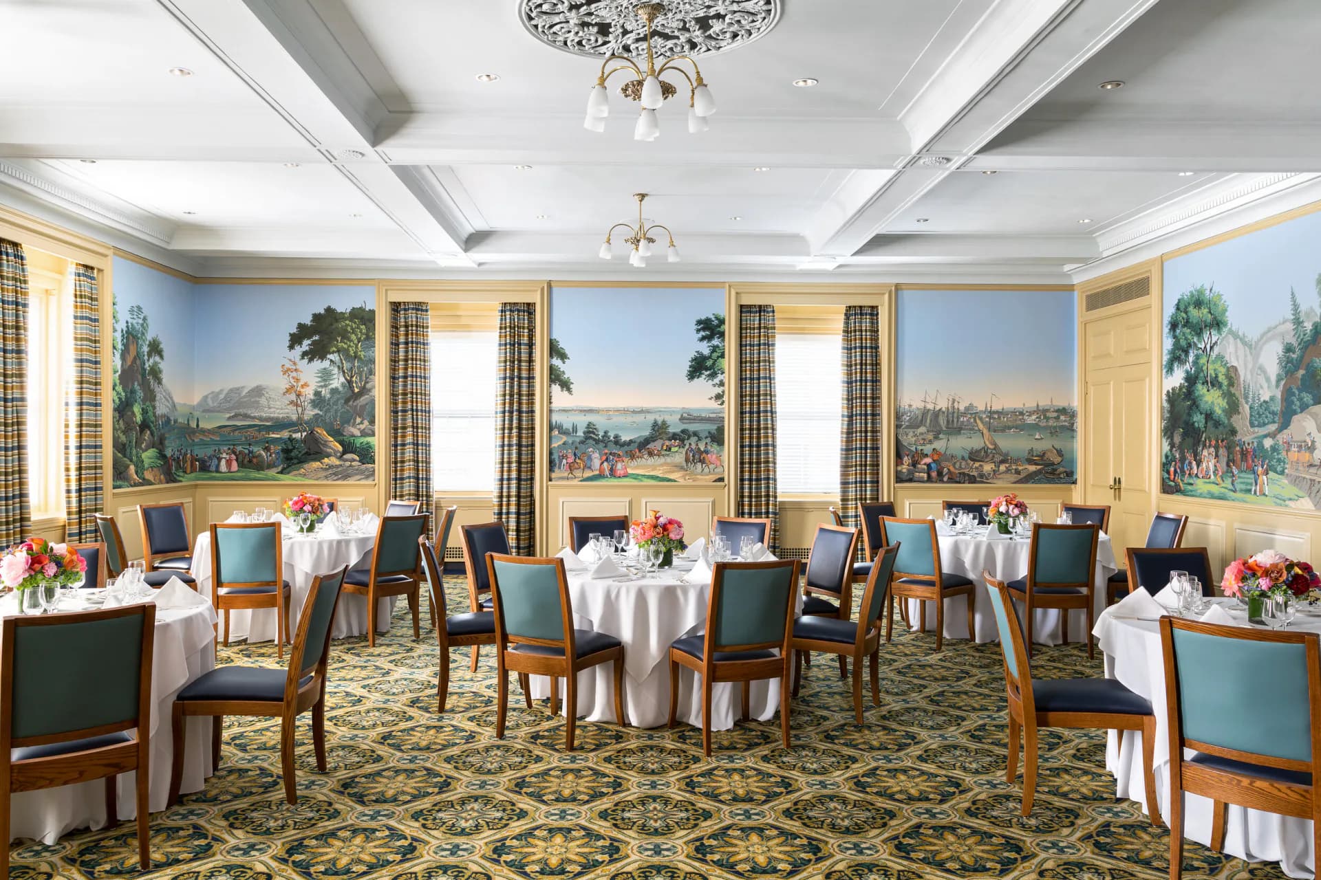 Elegant formal dining room with coffered ceiling, large-scale landscape murals, crisp white tablecloths, and blue-upholstered chairs by Eve Wildrick.