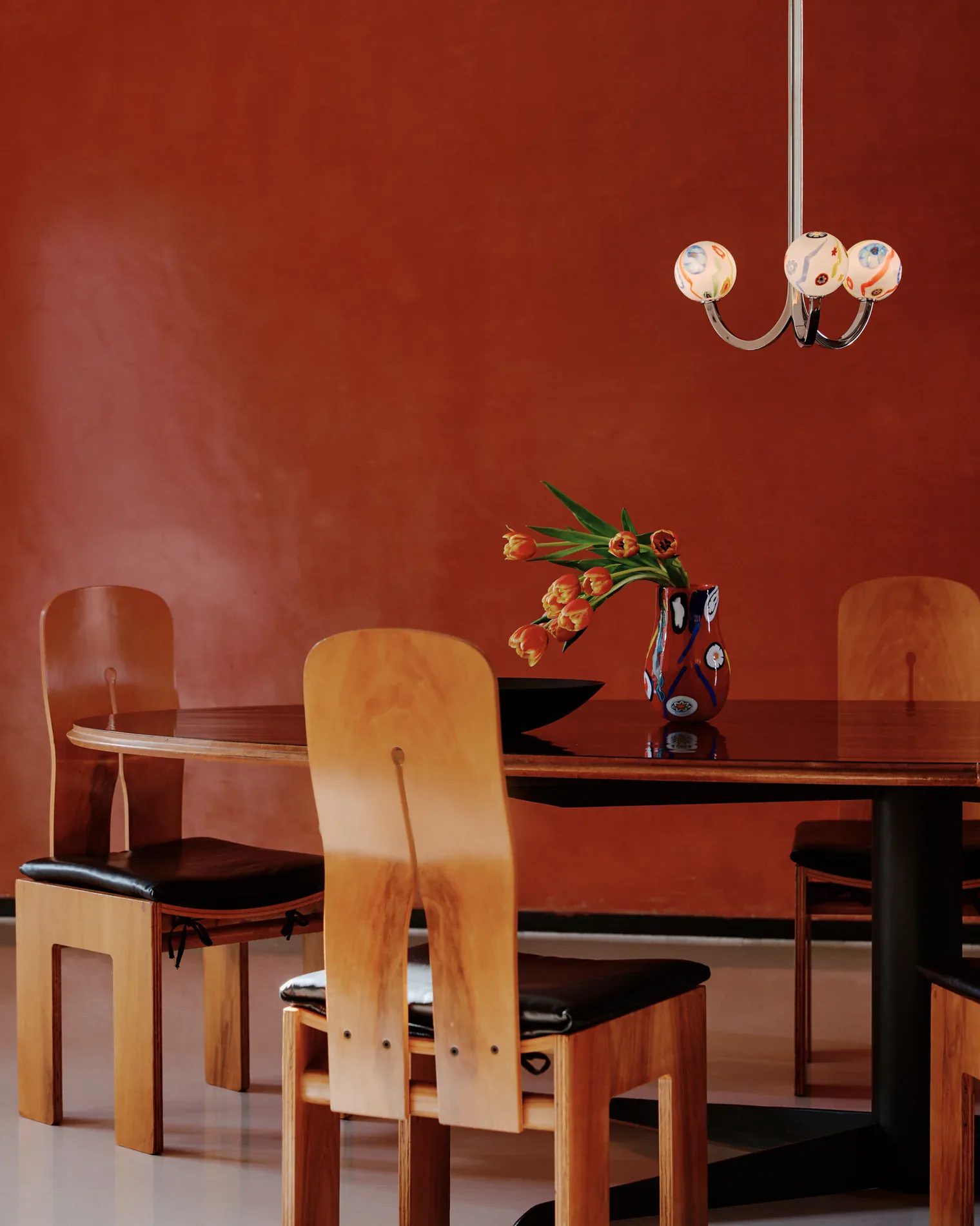 Modern dining room with warm terracotta walls, wooden chairs with black seats, black table, pendant lighting, and floral arrangement by Felicia Hung.