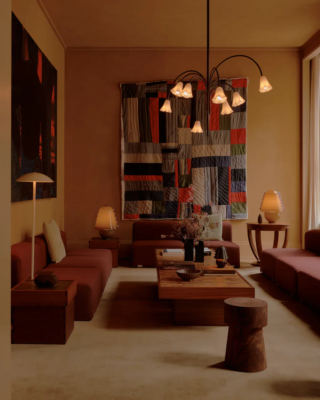 Warm-toned living room with red and burgundy upholstered seating, wooden coffee table, patterned textile wall art, and sculptural pendant lighting by Felicia Hung.