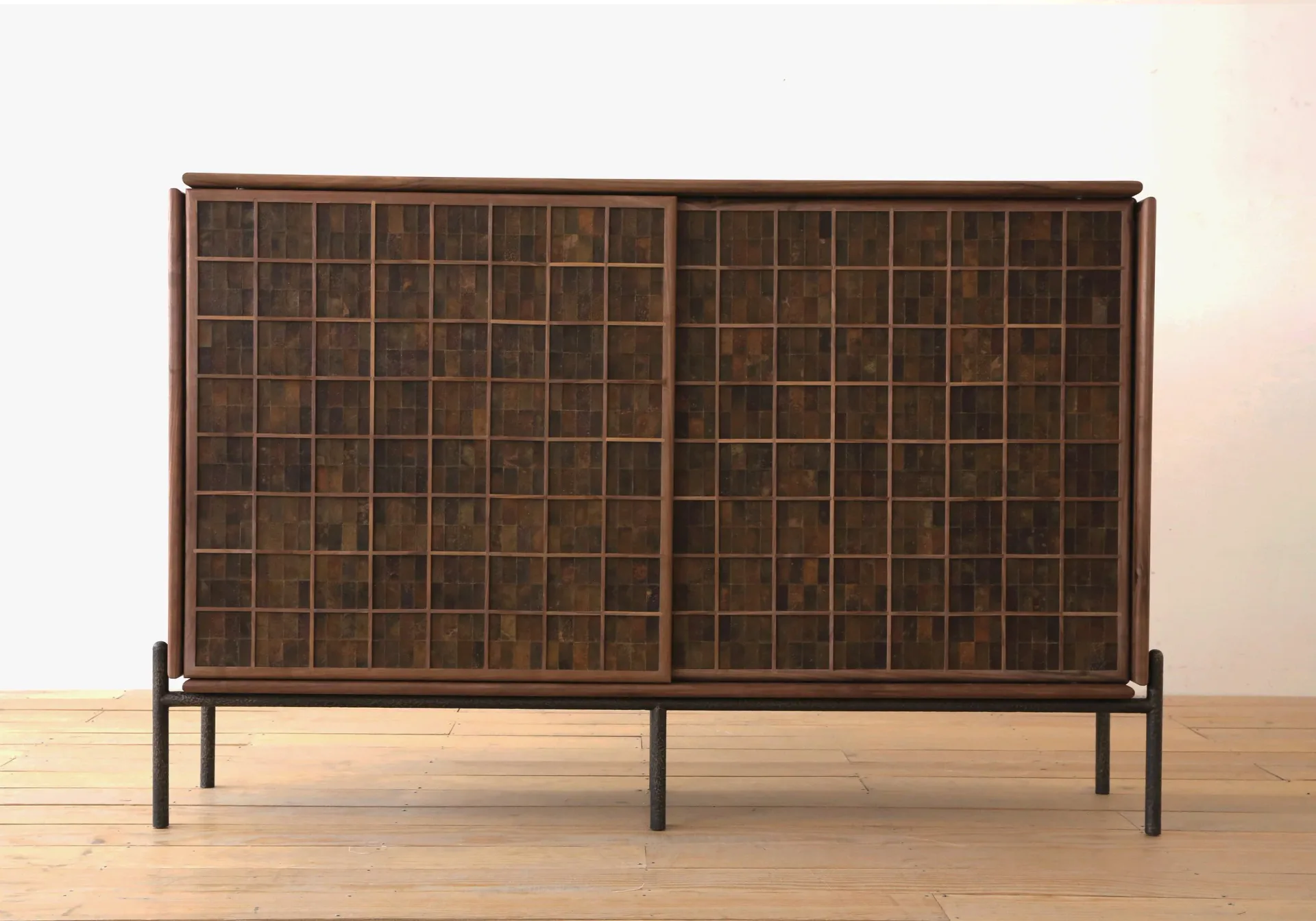 Dark wood cabinet with grid-patterned glass doors and metal frame legs on a light wooden floor by Fernando Laposse.