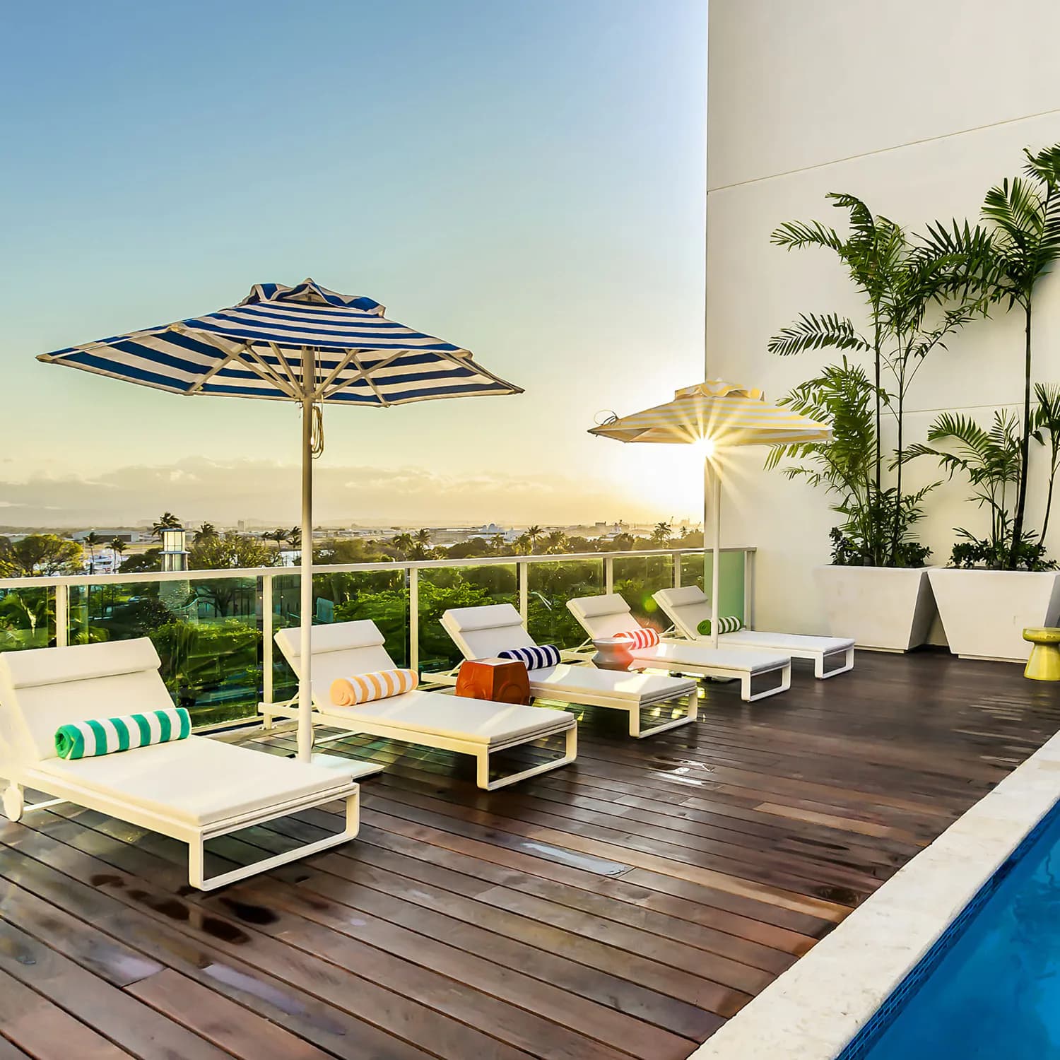 Rooftop terrace with striped umbrellas, white loungers with colorful cushions, wooden deck, climbing greenery, and city views by Fernando Rodriguez.