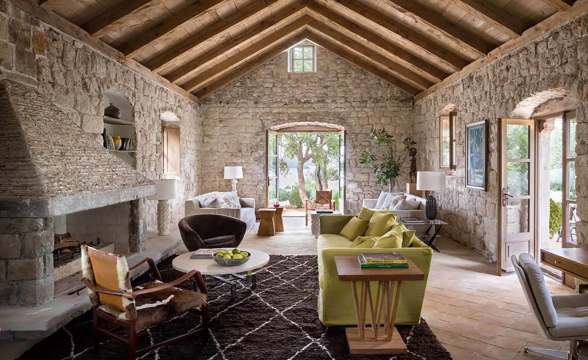 Stone residential interior with vaulted wooden ceiling, featuring chartreuse seating, fireplace, geometric rug, and natural light by Fiona Mueller.