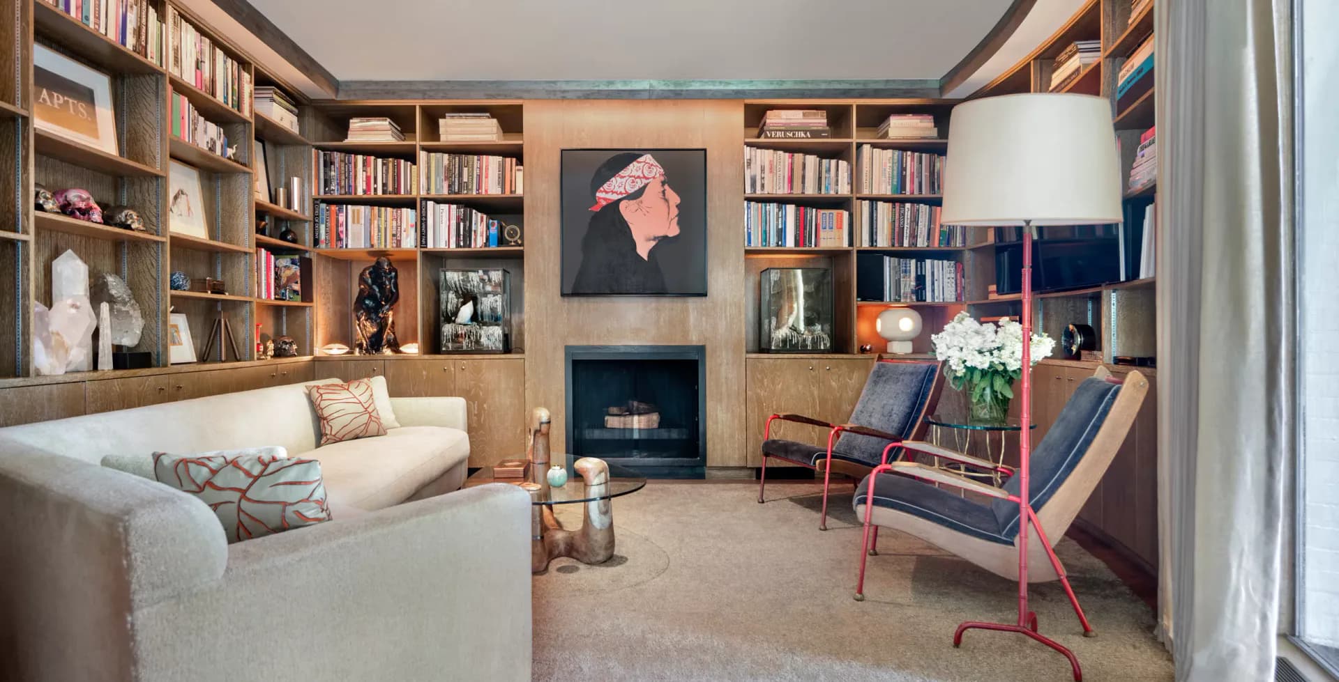 Warm-toned residential library featuring built-in wood shelving, cream sofa, fireplace, desk workspace, and mid-century modern furniture by Fiona Mueller.