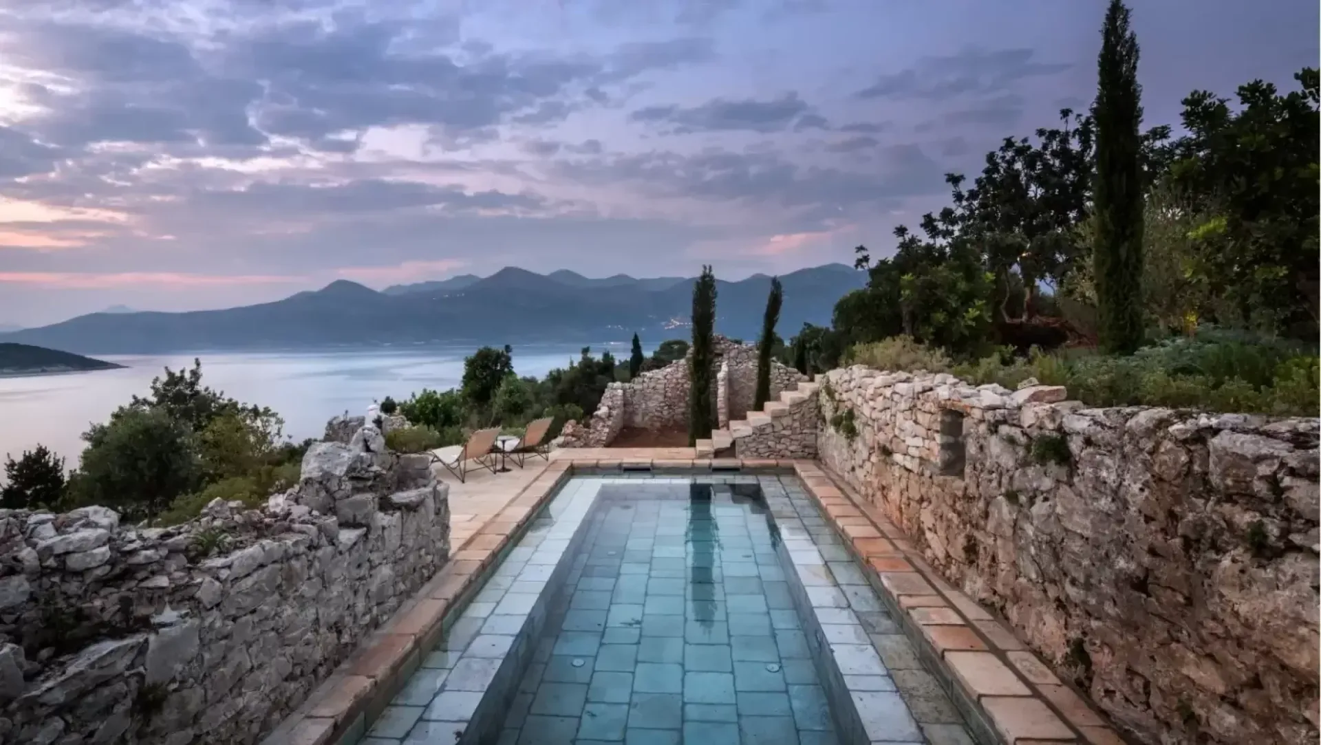 Infinity pool with teal tiles overlooking a Mediterranean coastal landscape with mountains and cypress trees at dusk, by Fiona Mueller.