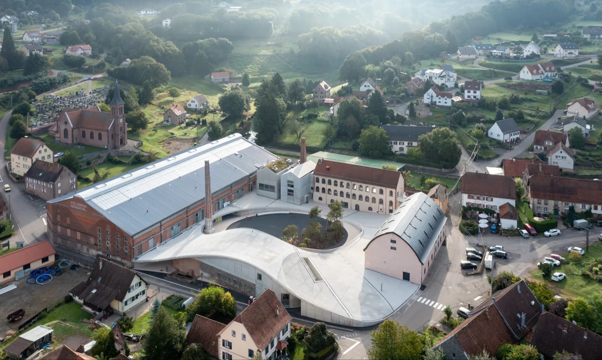 Aerial view of a modern institutional building with curved white roof forms and brick facades set within a green valley landscape, designed by Florian Idenburg.