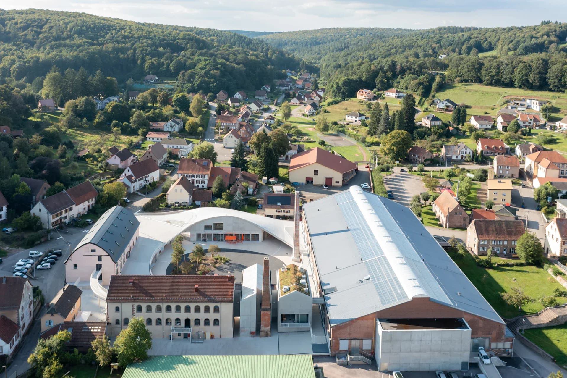 Aerial view of a contemporary institutional building with curved metal roofing integrated into a residential hillside village landscape by Florian Idenburg.