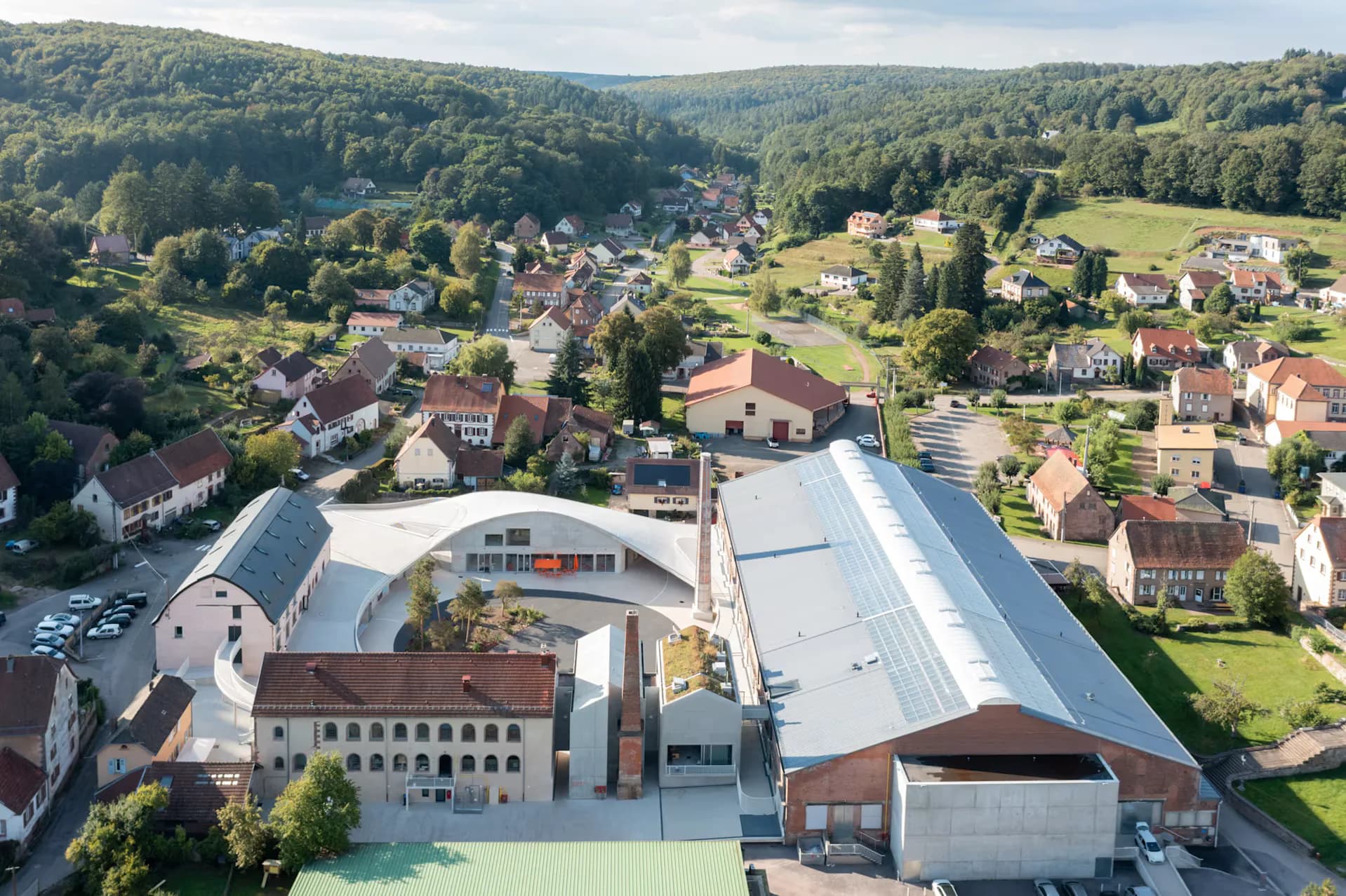 Aerial view of a modern institutional building with white metal roof and brick facade set in a verdant valley village landscape by Florian Idenburg.
