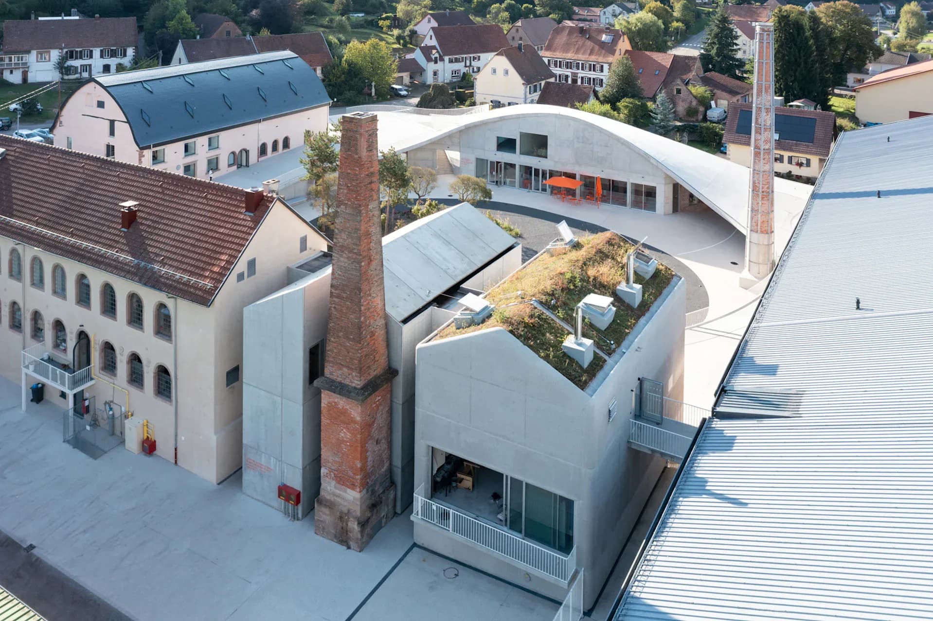 Aerial view of a modern white building with a preserved brick chimney, green roof terrace, and integrated courtyard, surrounded by traditional residential architecture by Florian Idenburg.