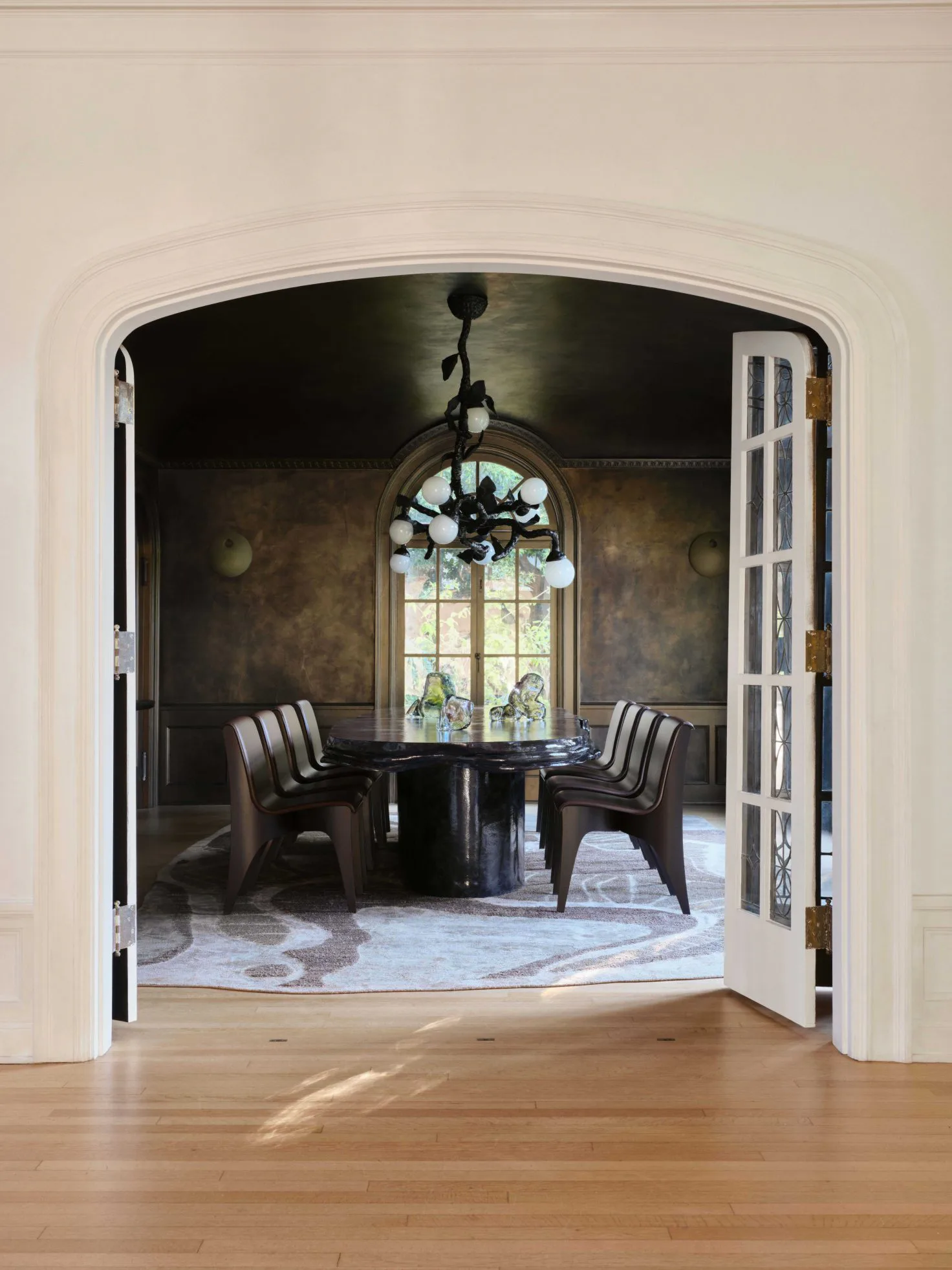 Elegant dining room with dark walls, black round table, striped chairs, and arched doorway with stained glass window by Floris Wubben.