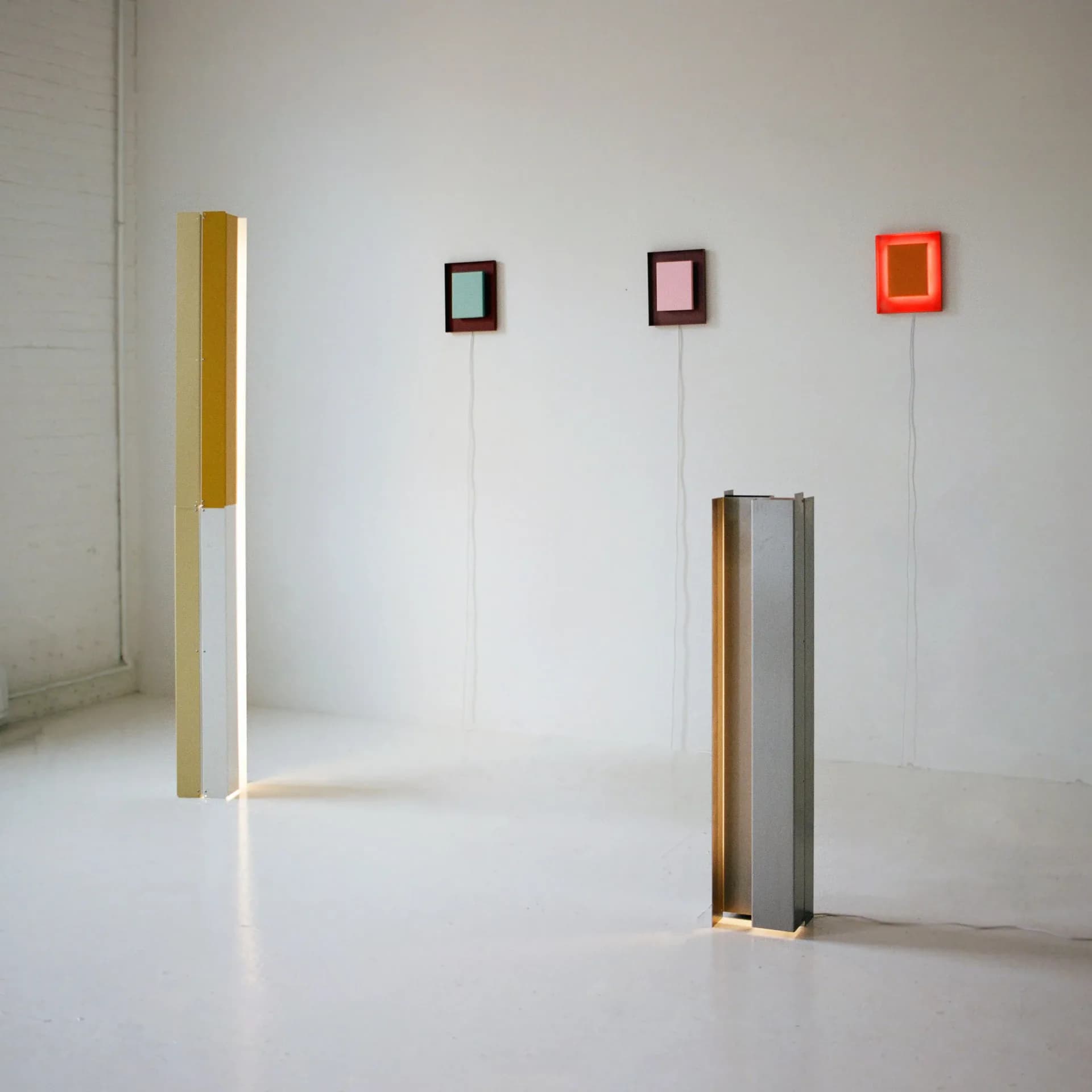 Minimalist installation with suspended geometric forms in muted tones—golden cylinder, brown and gray rectangular volumes, and small colored cubes in a white gallery space by Ford Bostwick.