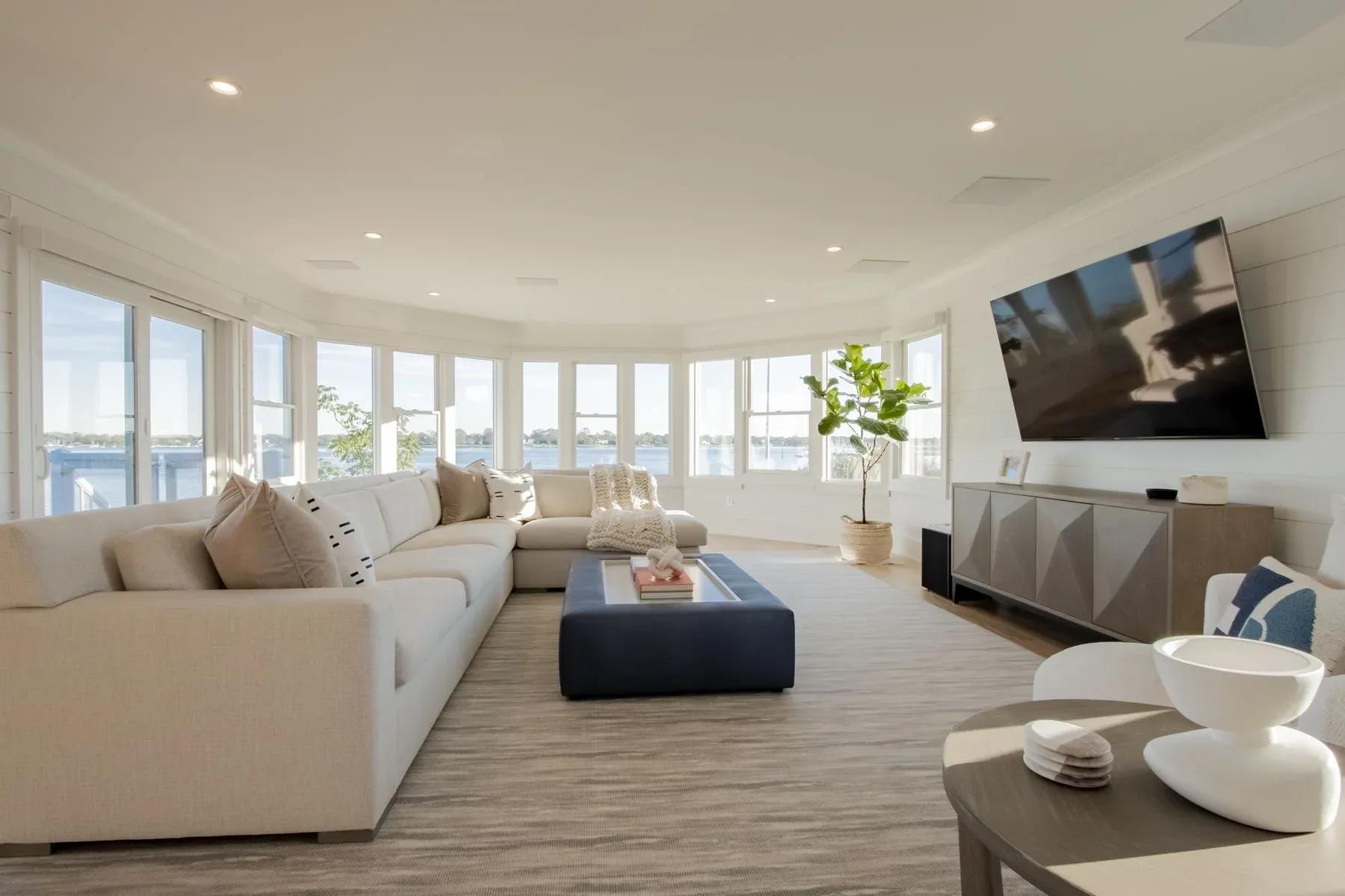 Bright modern living room with curved white sectional, black ottoman, extensive waterfront windows, and mounted TV, designed by Fotini Levy.