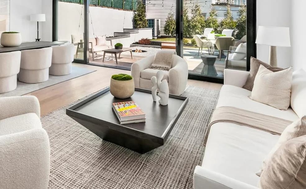 Minimalist living room with cream upholstered seating, black low coffee table, neutral area rug, and expansive glass doors opening to a patio by Francesca B Bova.